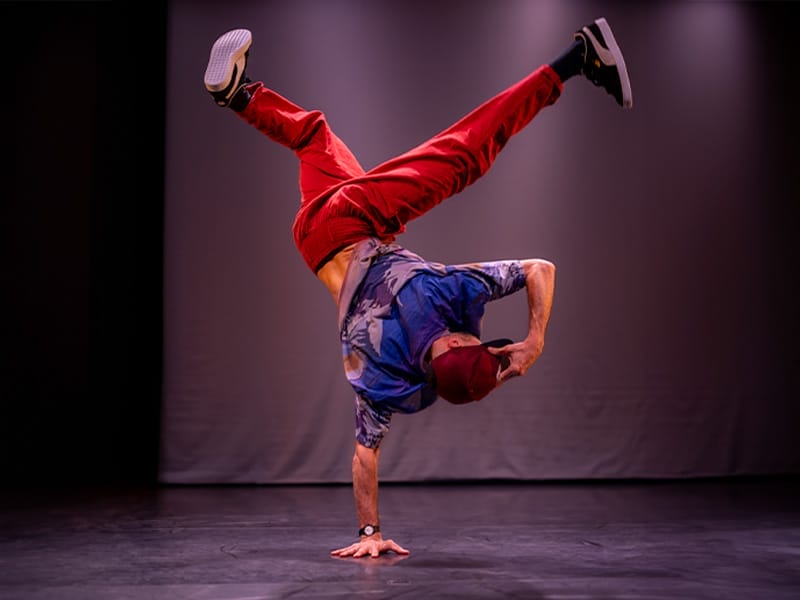 A man is breakdancing on a dark stage, with his back to the camera he poses in a one-handed handstand, with legs outstretched.