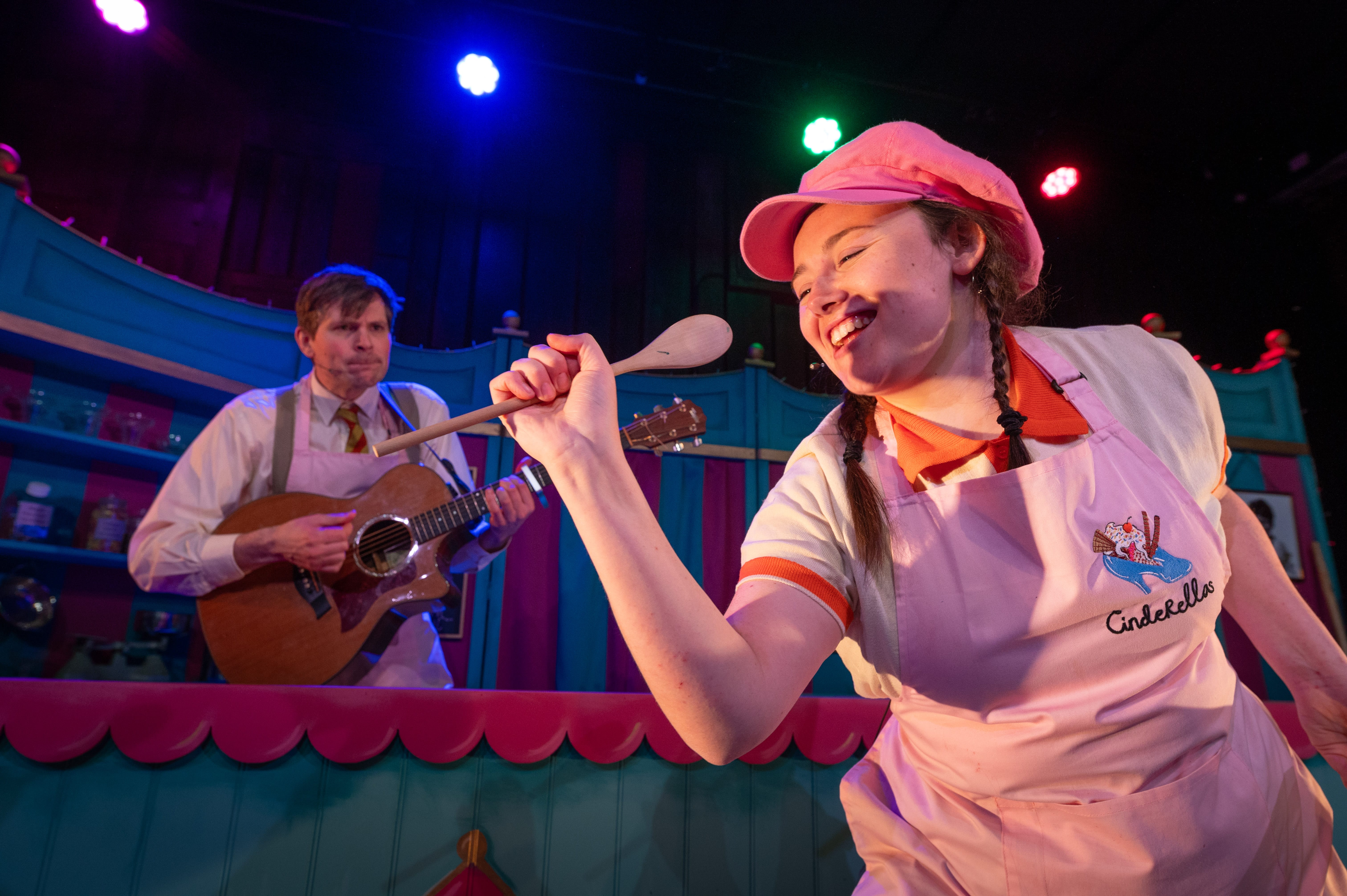 A girl wearing a pink hat sings into a wooden spoon, and a man plays the guitar in the background.