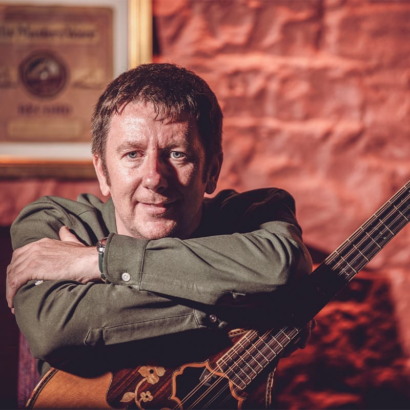 A man with short brown hair leans on his bouzouki guitar.