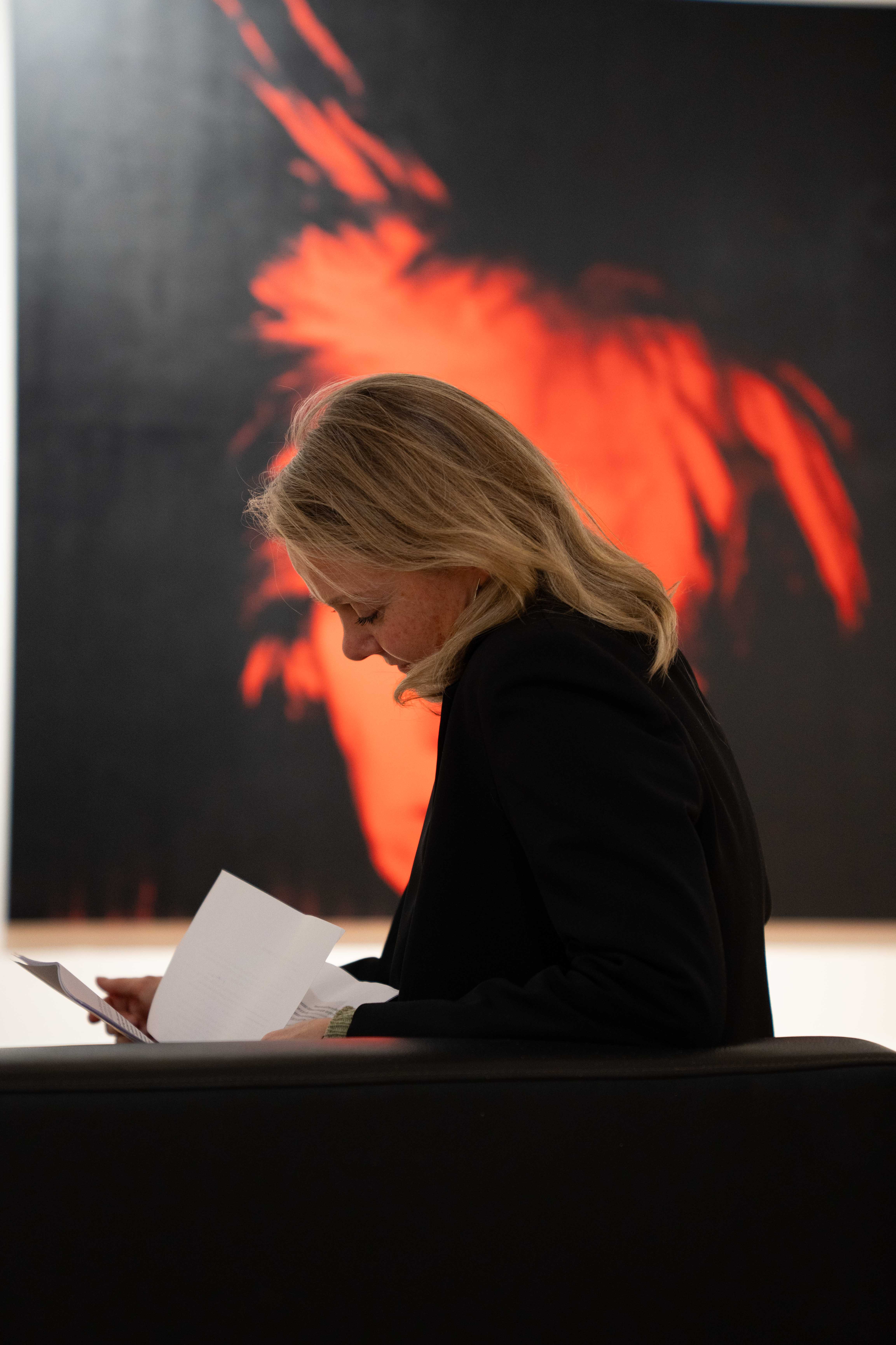 A woman with blond hair sits side on, reading notes in front of Andy Warhol artwork.