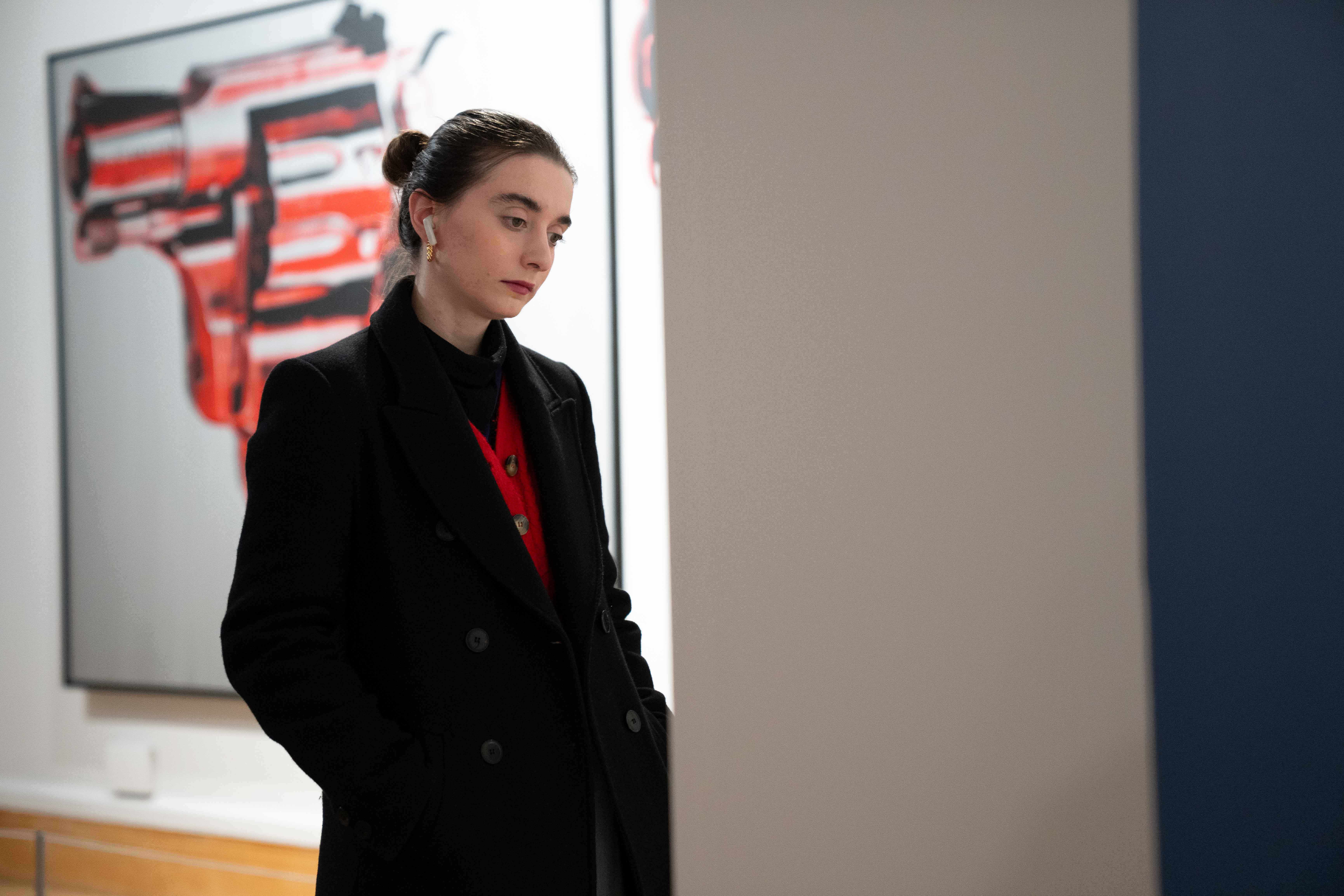 A young woman wearing a long dark coat admires artwork in a gallery.