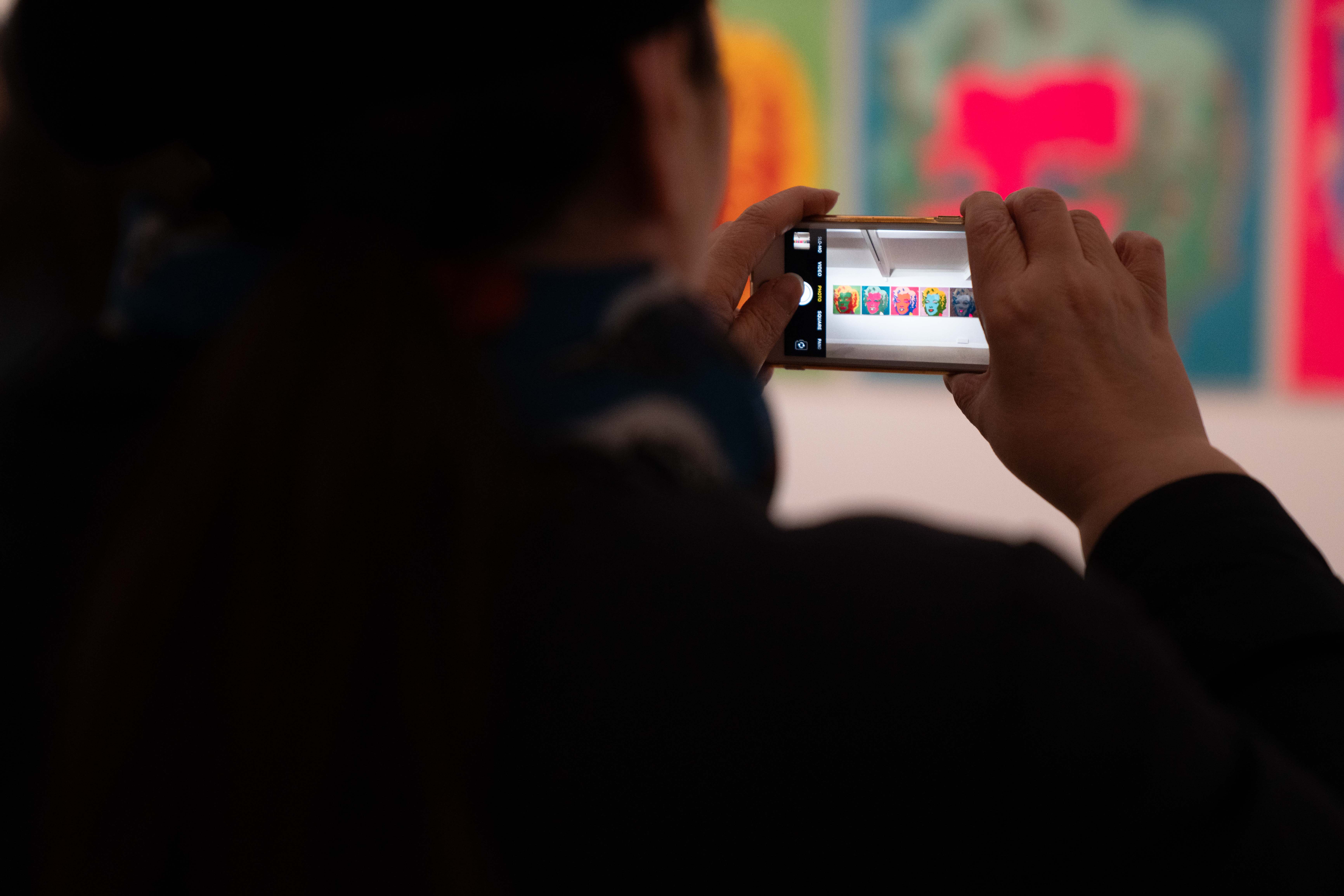 Looking over the shoulder of someone taking a photo of Andy Warhol artwork with their phone.