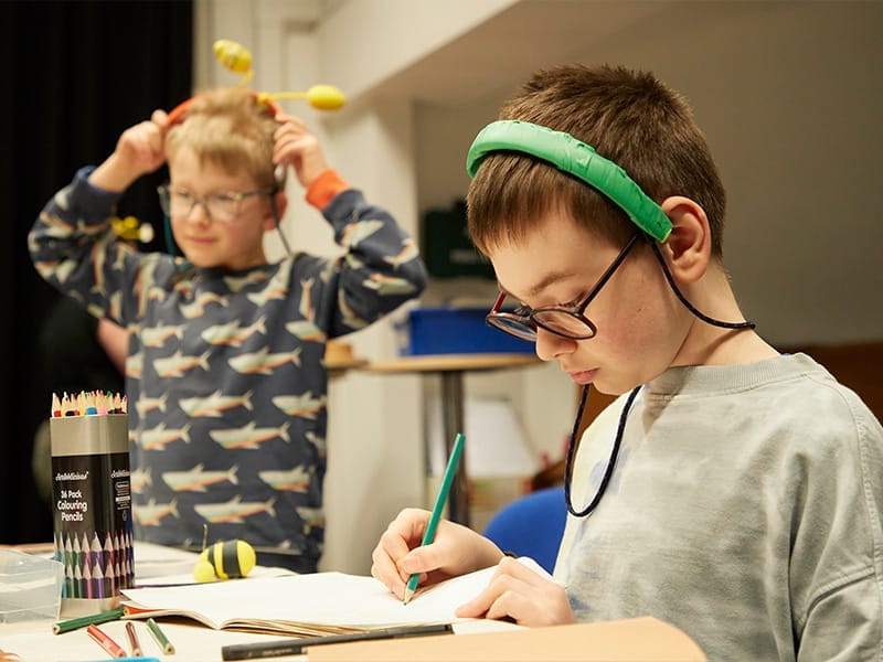 One boy draws in a notepad with a green pencil, whilst the other shows off his handmade headband.