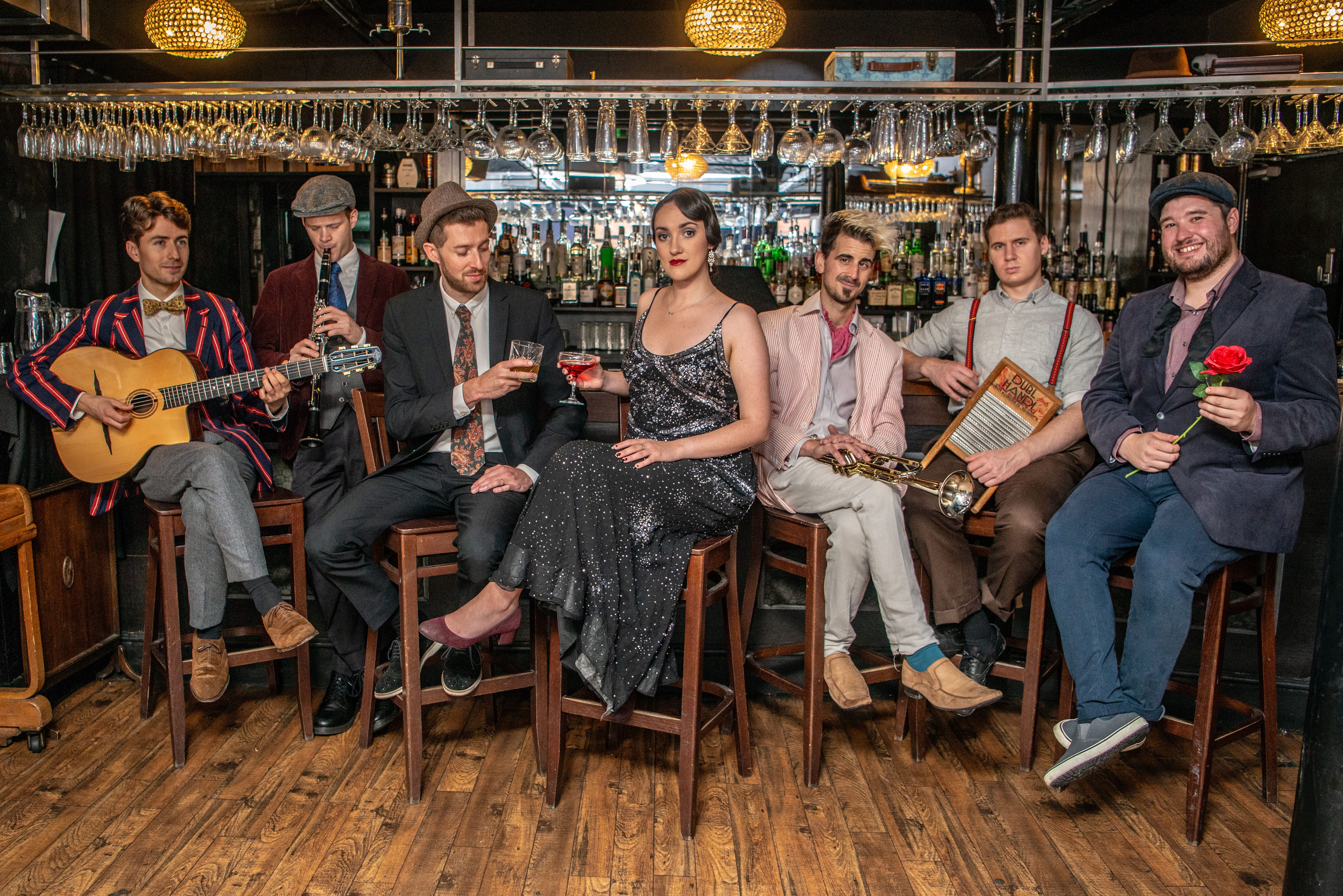 A jazz group, made up of 7 musicians, sit at a cocktail bar, holding drinks or their instruments.