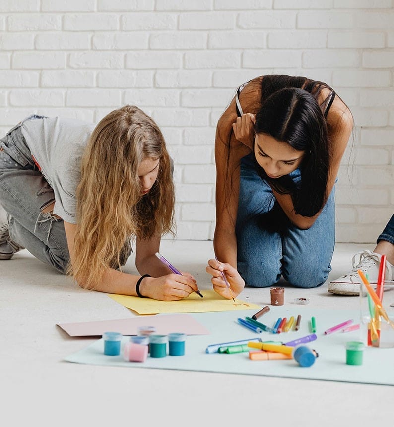 Two teen girls kneel on the floor whilst painting.