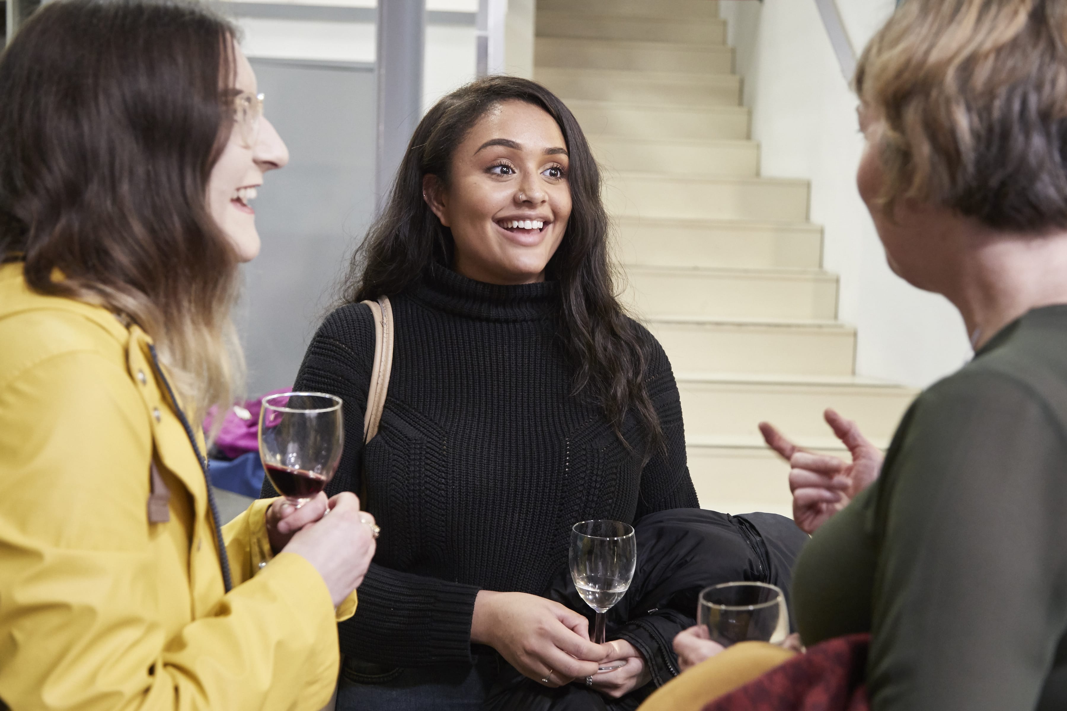 Three people holding glasses of wine in their hands are talking to each other with a woman in the middle of the picture in the focus, she is smiling and pays attentions to the what the other person is saying.