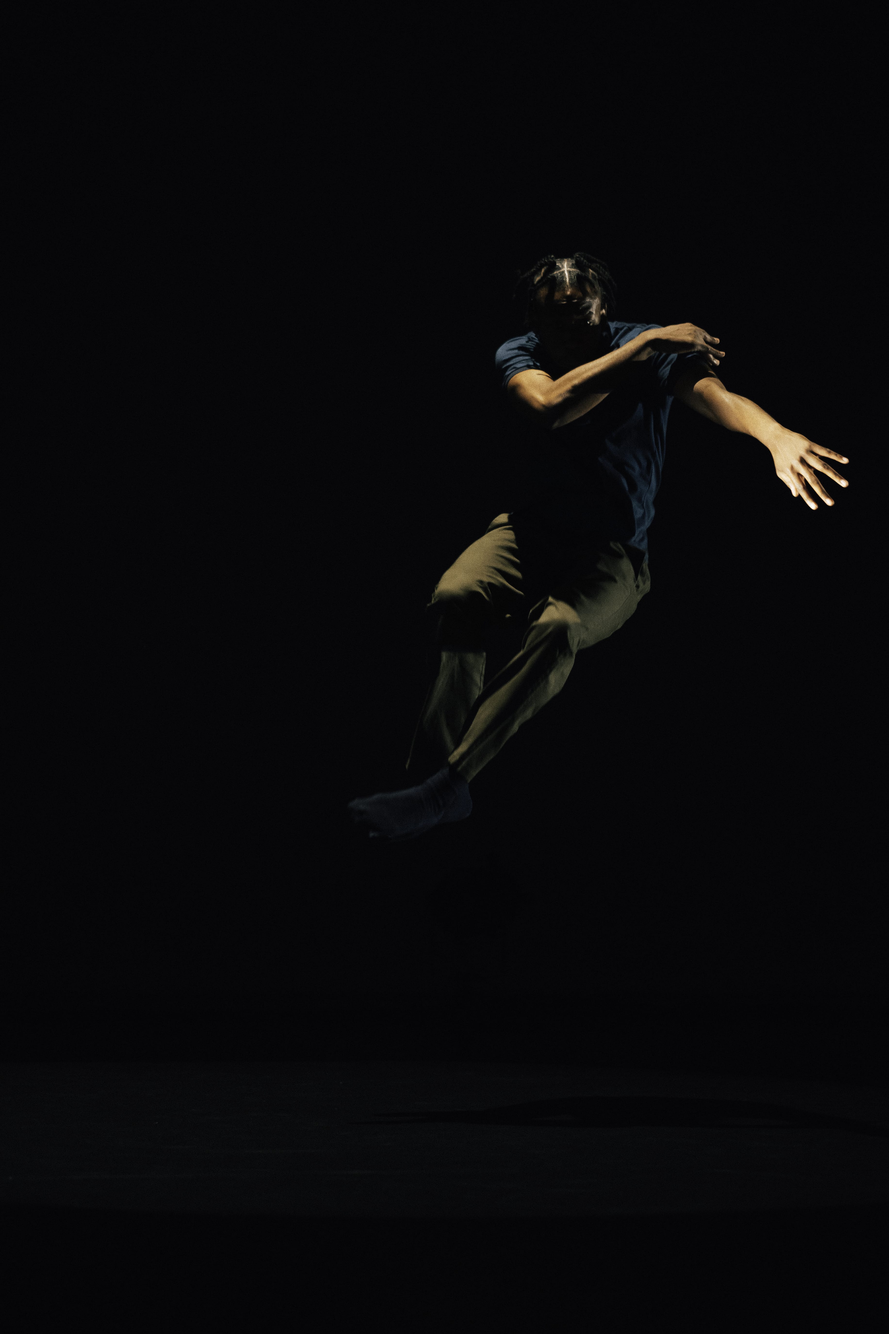A dancer wearing a blue t-shirt and pale trousers, jumps, folding his arms in a dark space.