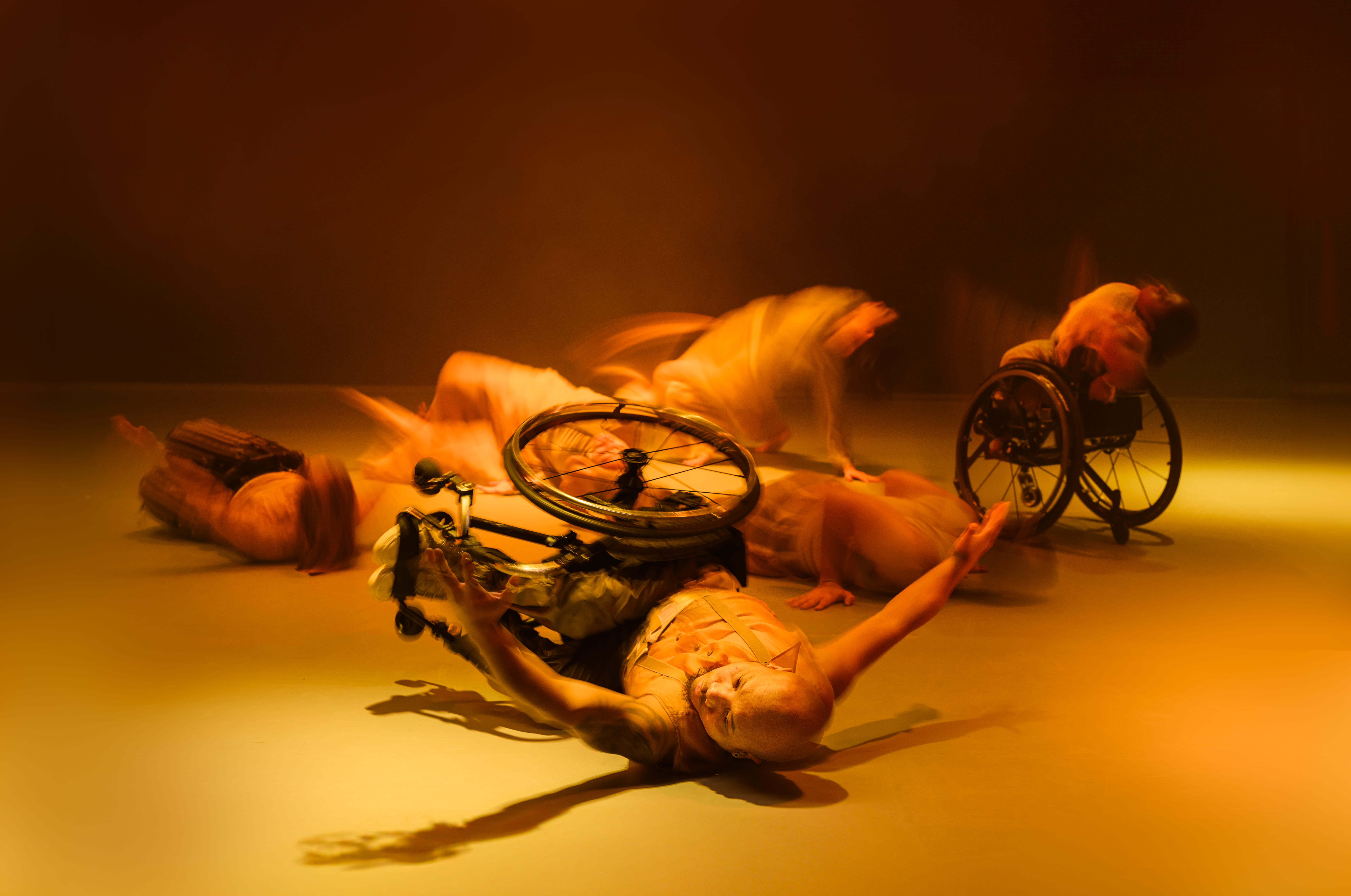 An action shot of a dance scene. A male wheelchair dancer with brown skin, is lying sideways on a light coloured floor. His wheelchair is still strapped on his waist, lying on the floor with him. His arms are raised off the floor, and he is looking intently at his left hand. He is in control. Other dancers are in the background in blurry motion shots, all on the floor making shapes that are reflective of the wheelchair dancer's pose but all different in their own ways. A female wheelchair dancer with dark skin and black hair is blurry but more visible to the right of the image. She is facing away from the camera, tipping backwards, her head going over the back rest of her chair. The image has a golden colour with atmospheric haze and black backdrop. The dancers are all in stylised tight costumes that has a golden burnished colour. Photography by Chris Parkes