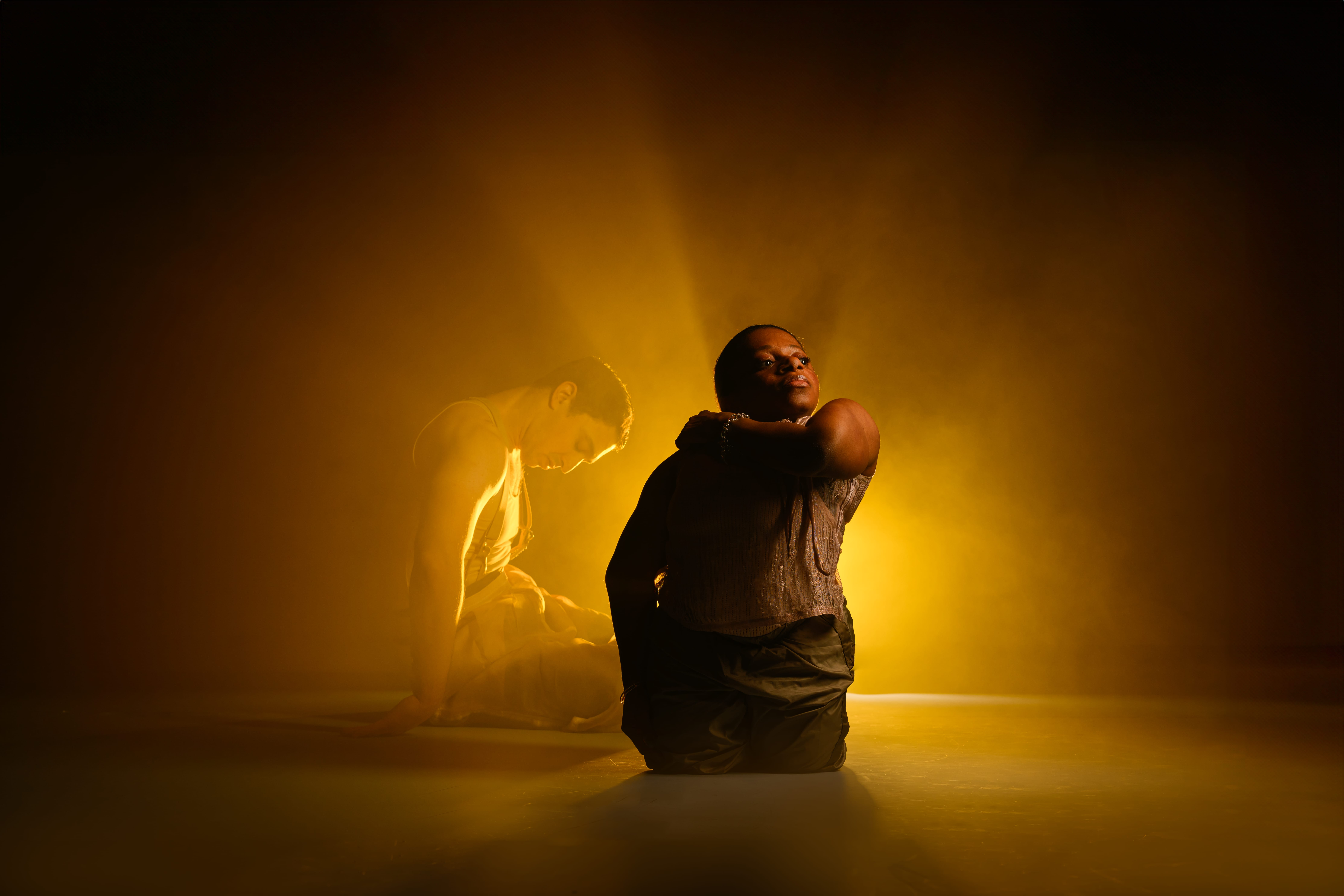 A golden, burnished photo of dancers Mo and Christian during a duet. Mo, a short-statured Disabled Black woman, kneels up and proudly holds her right arm across her chest to her heart, facing front she gazes out and across to the left of the image, her chin raised. Hidden slightly behind her, Christian, a tall non-disabled white man also kneels but faces his body to the right, but he curls himself downwards, his face gazing the floor. The image is hazy and back lit, having a black and golden background. Photography by Chris Parkes