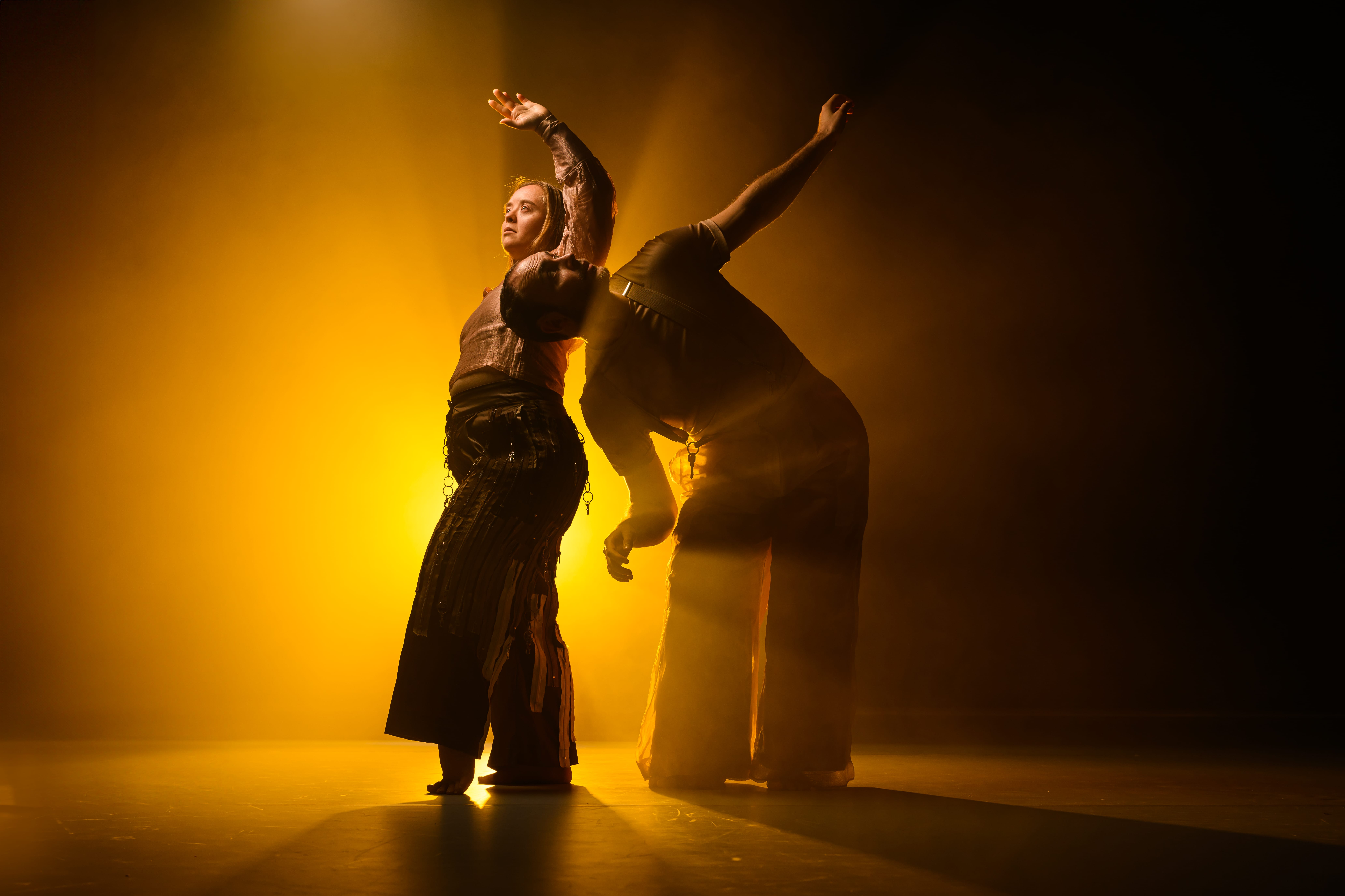 A female caucasian dancer with Down's Syndrome is standing with firm strong gaze cast upwards. Her left hand is raised above her head as if answering to a calling. Her left foot is raised on her toes, her left softly bent at the knee. She is wearing dark brown trousers with zips and fasteners stitched on all over like in a punk aesthetic. She is wearing a crumpled, peach coloured blouse. Beneath her raised arm alongside her torso, is the head of caucasian male dancer. He is bent forwards at the hip, his head directly under her armpit looking up at her raised hand. His left arm is raised, as if left behind him as he bent forward. He is almost completely silhouetted. There is a key dangling on his vaguely visible braces. Image by Chris Parkes.