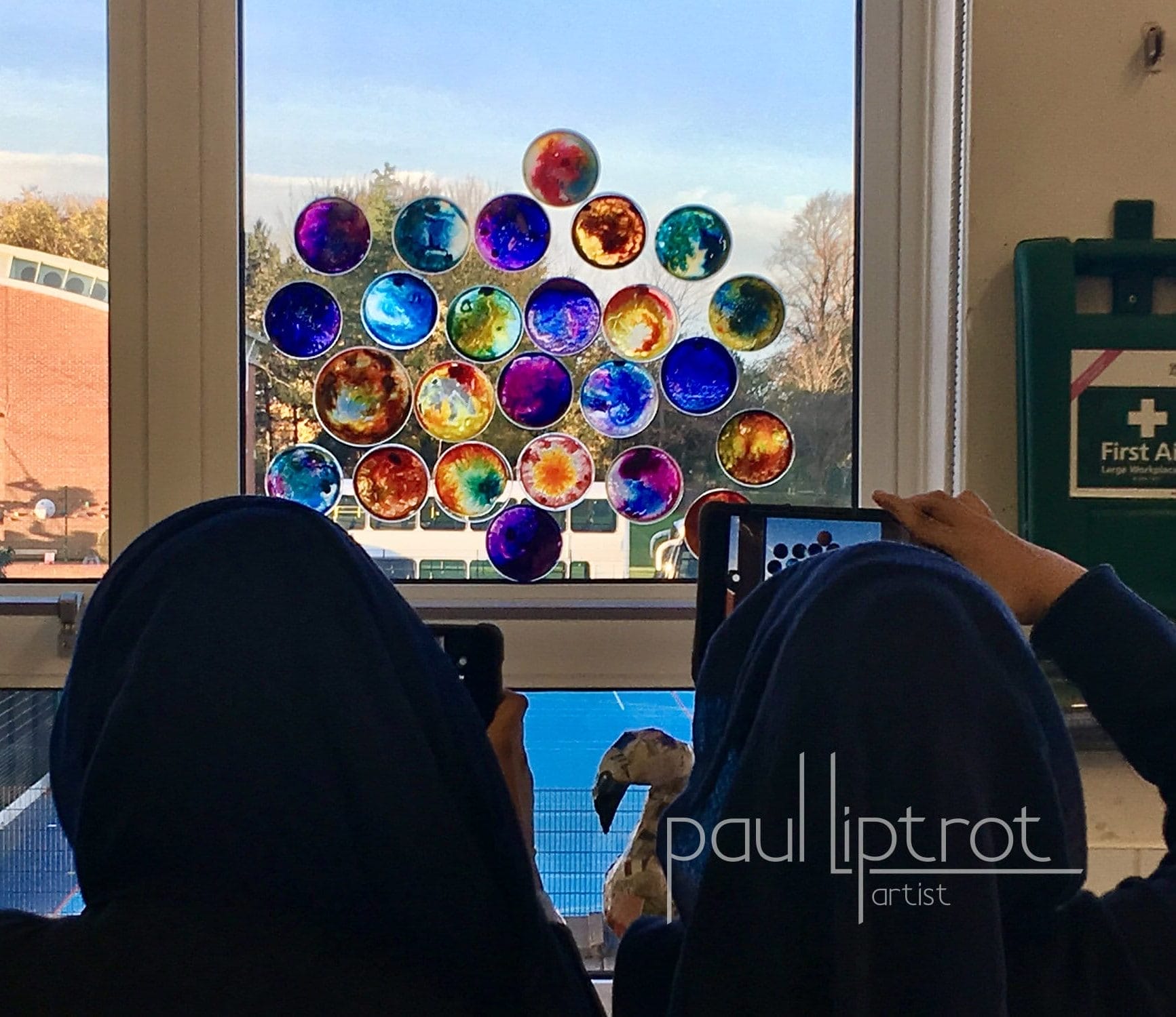 two girls wearing headscarfs look how the light comes in through colourful petri dishes placed at a window
