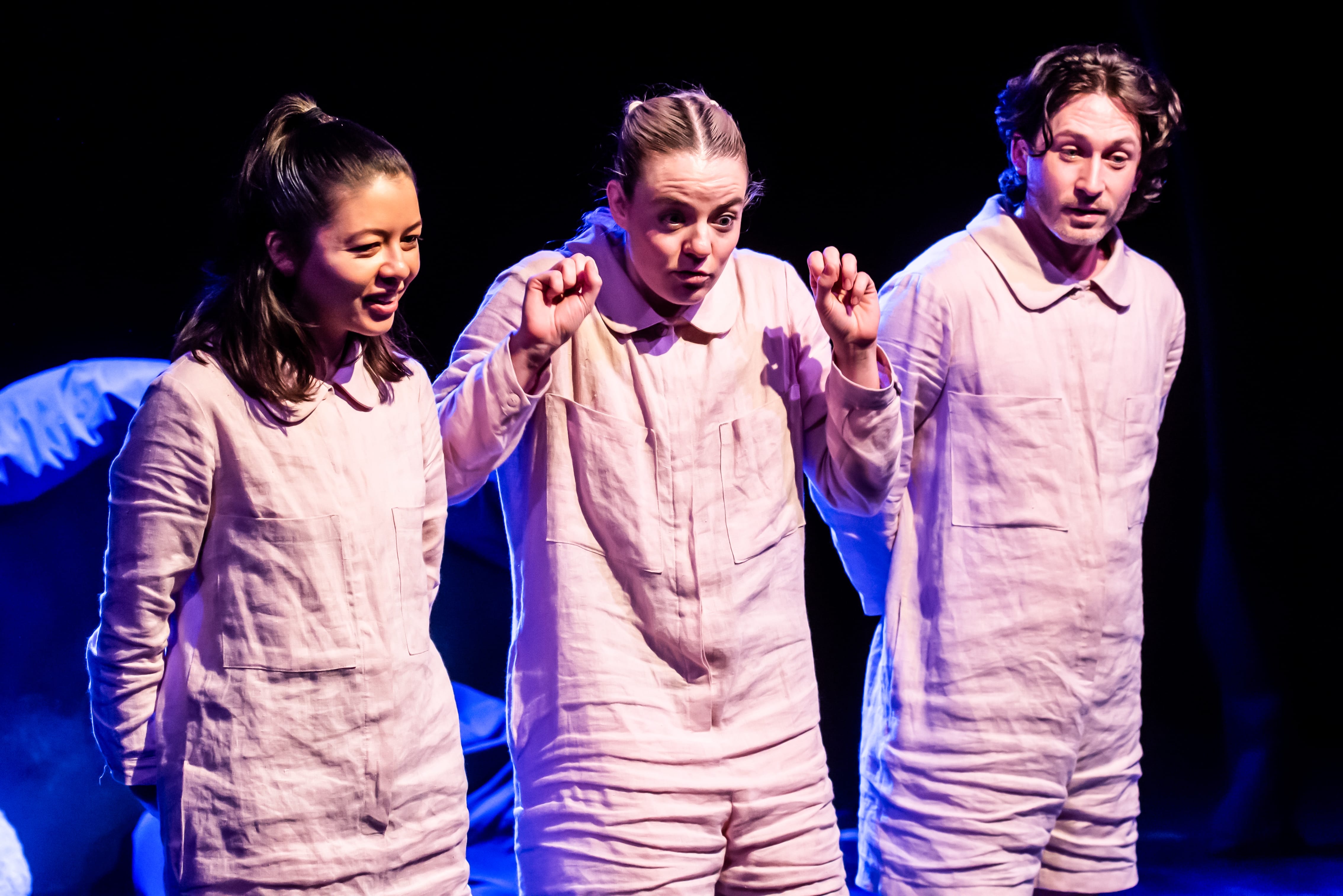 Three performers stand on stage, wearing beige linen jumpsuits.