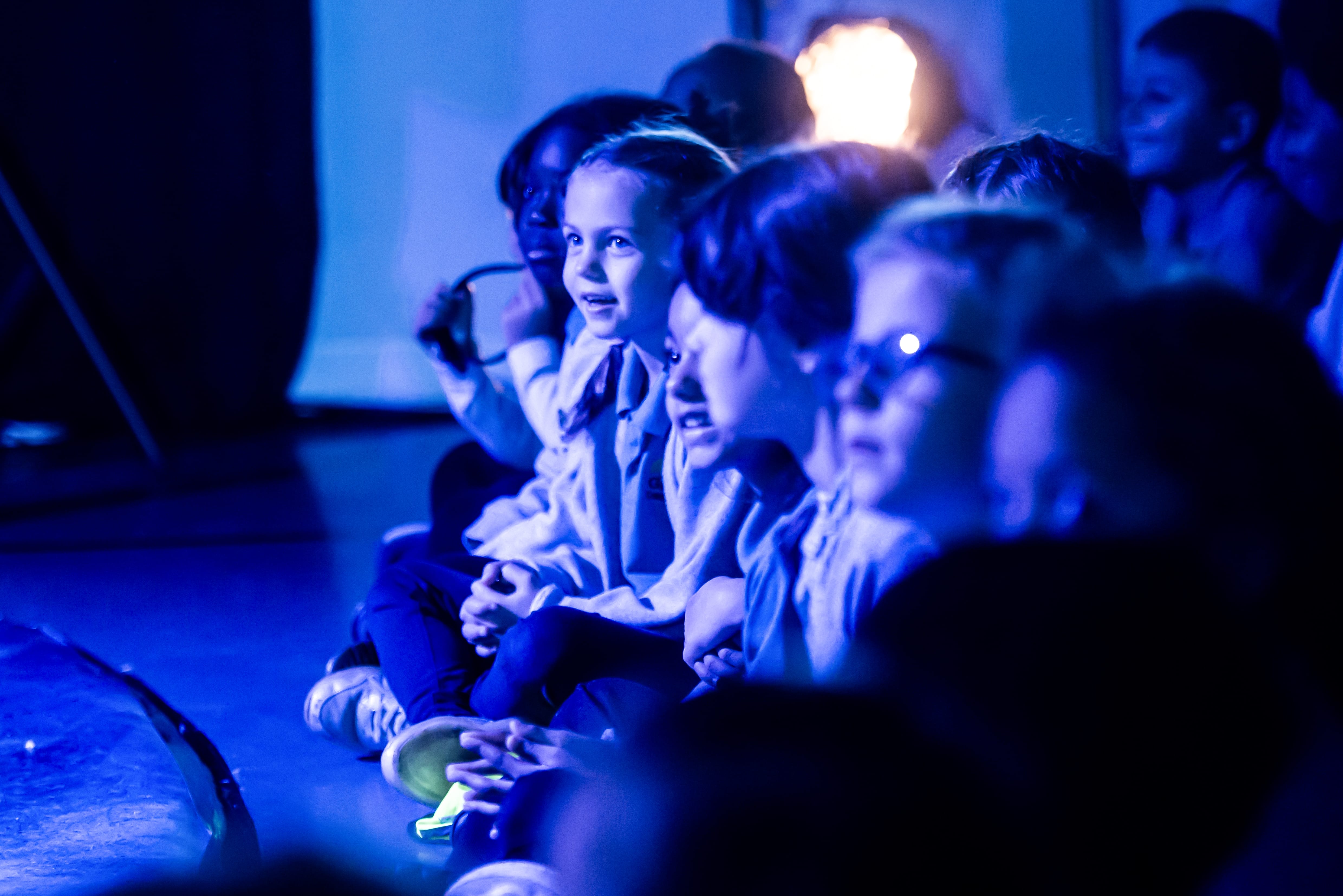 Children sit, under blue light, captivated by a performance on stage.