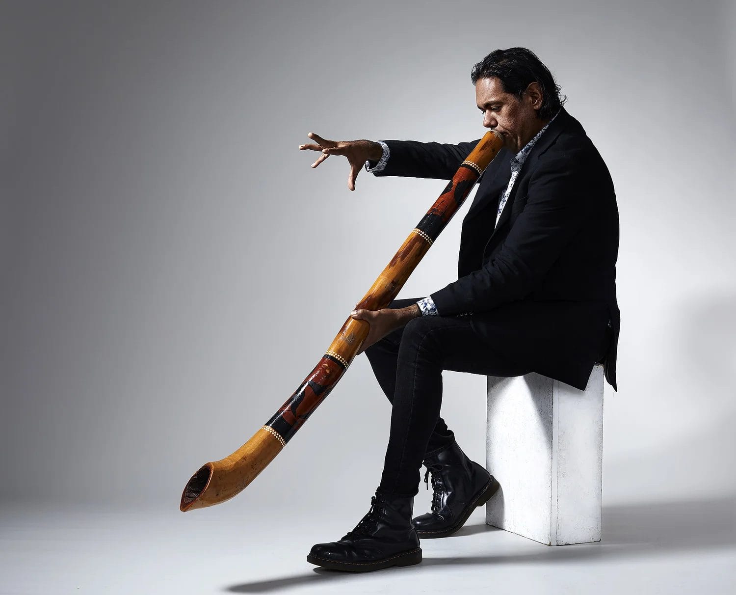 A man in a black suit, sits on a white box playing the didgeridoo