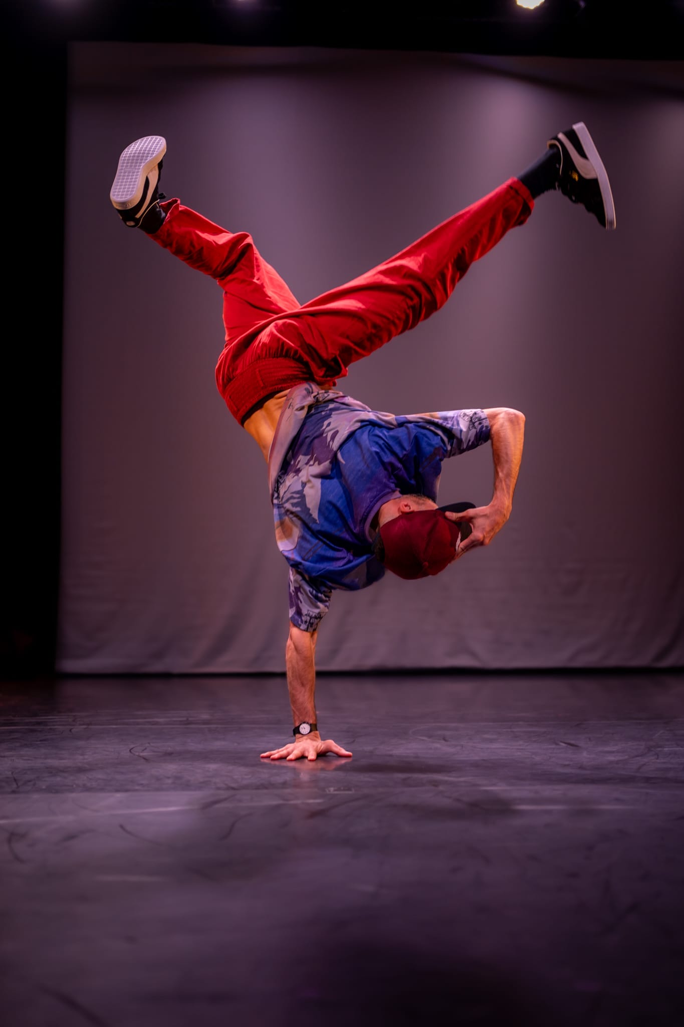 A photo of a man breakdancing on stage. He wears red joggers and a blue t-shirt and a cap. He stands on one hand.