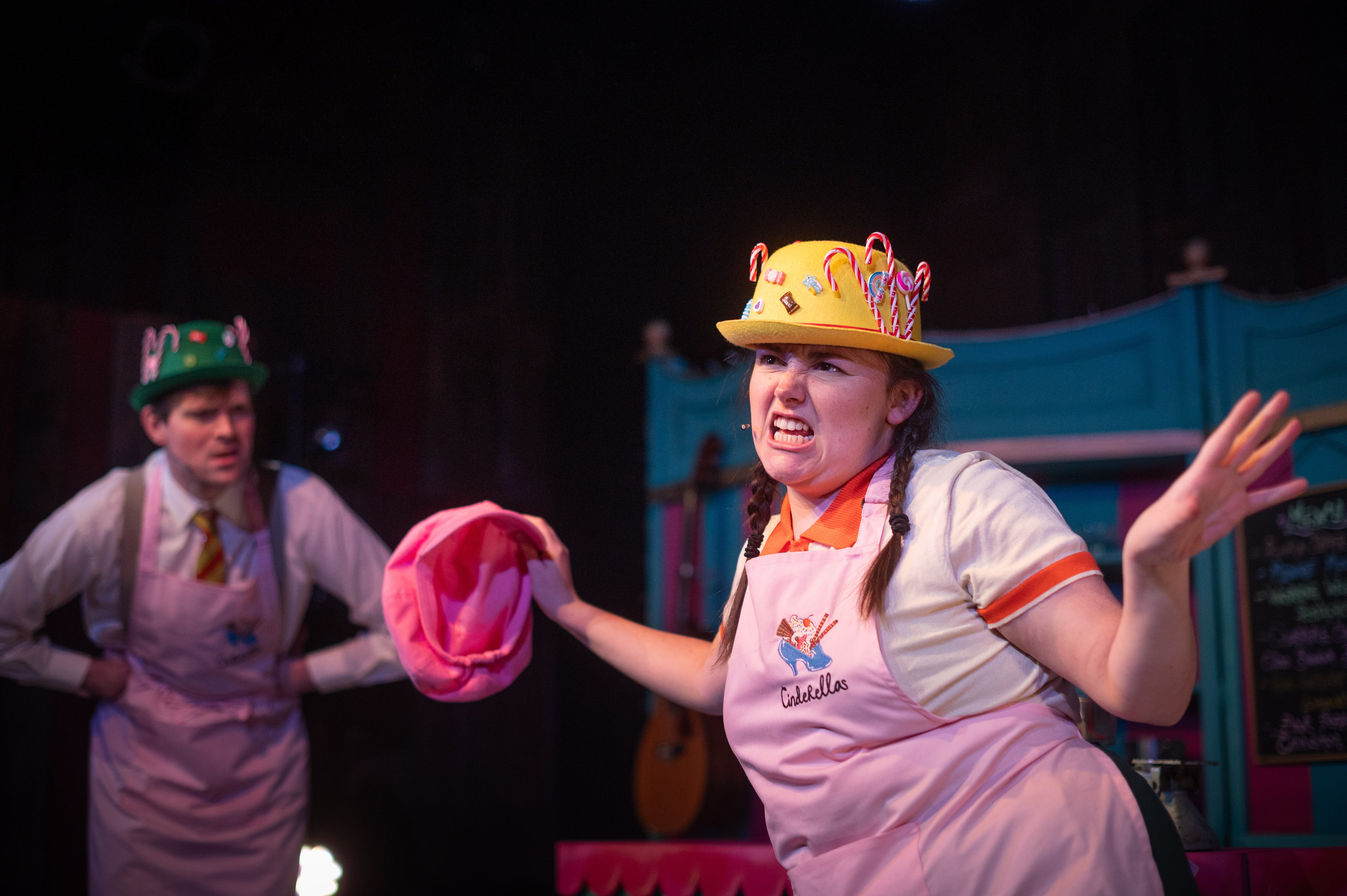 A woman with her brown hair in plaited pigtails wears a yellow hat and pink apron. Another performer stands in the background also wearing a pink apron.