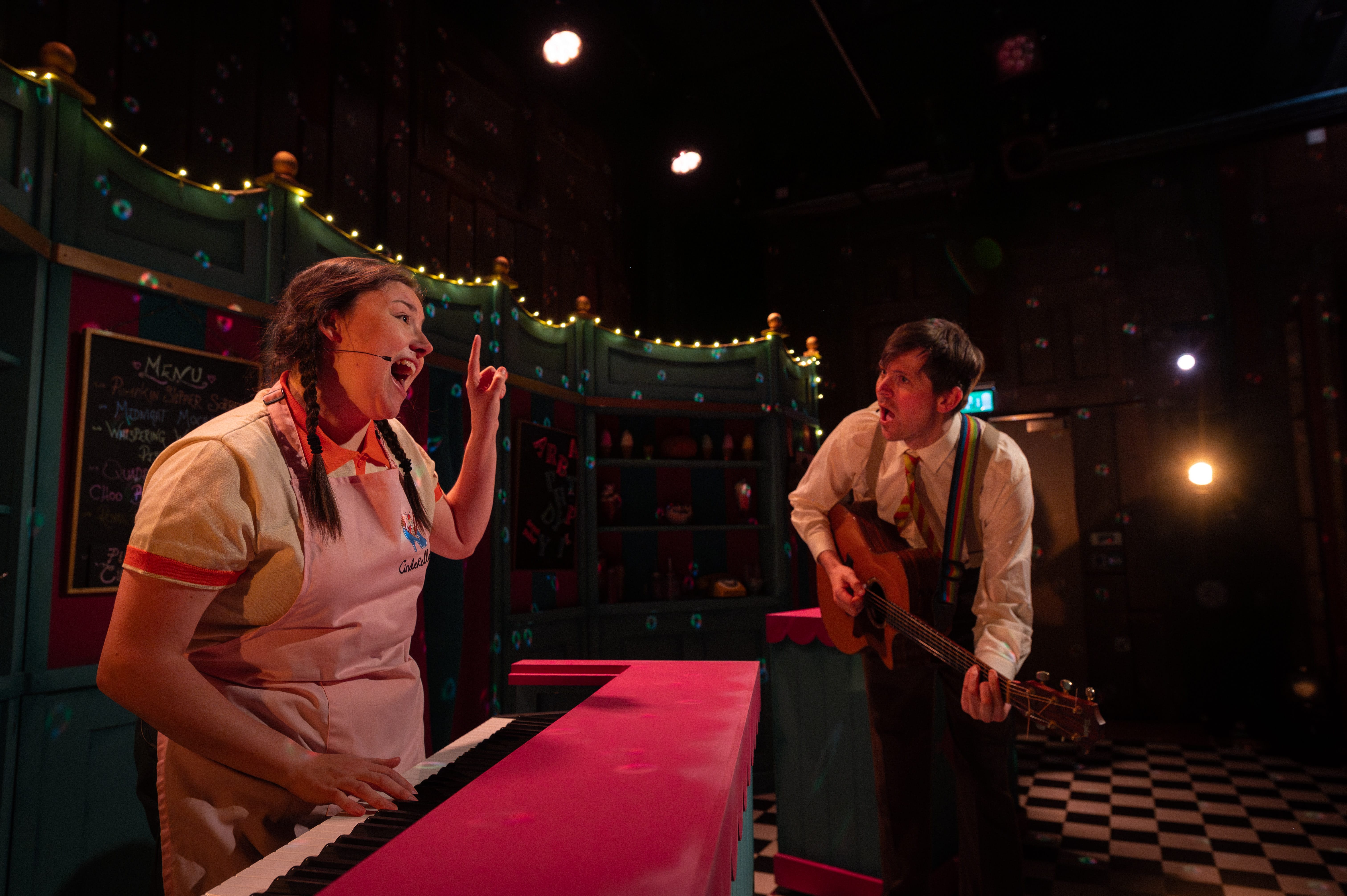 A woman stands playing a keyboard, with one finger in the air. Whilst a man wearing a white shirt and tie plays the guitar.