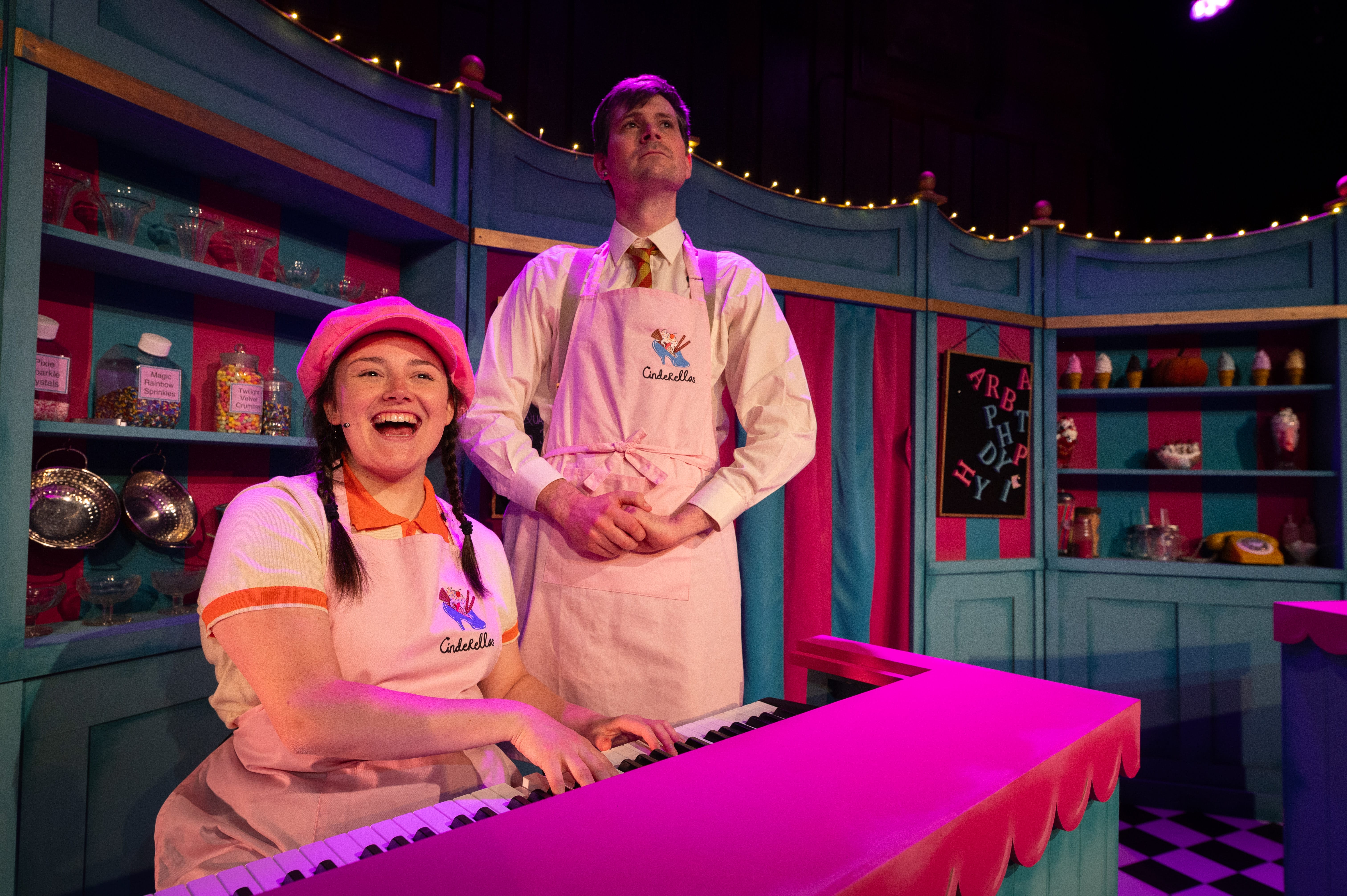 A woman wearing a bright pink hat and pink apron sits playing a keyboard. A man stands behind with his hands held together, also wearing a pink apron and looking out to the audience.