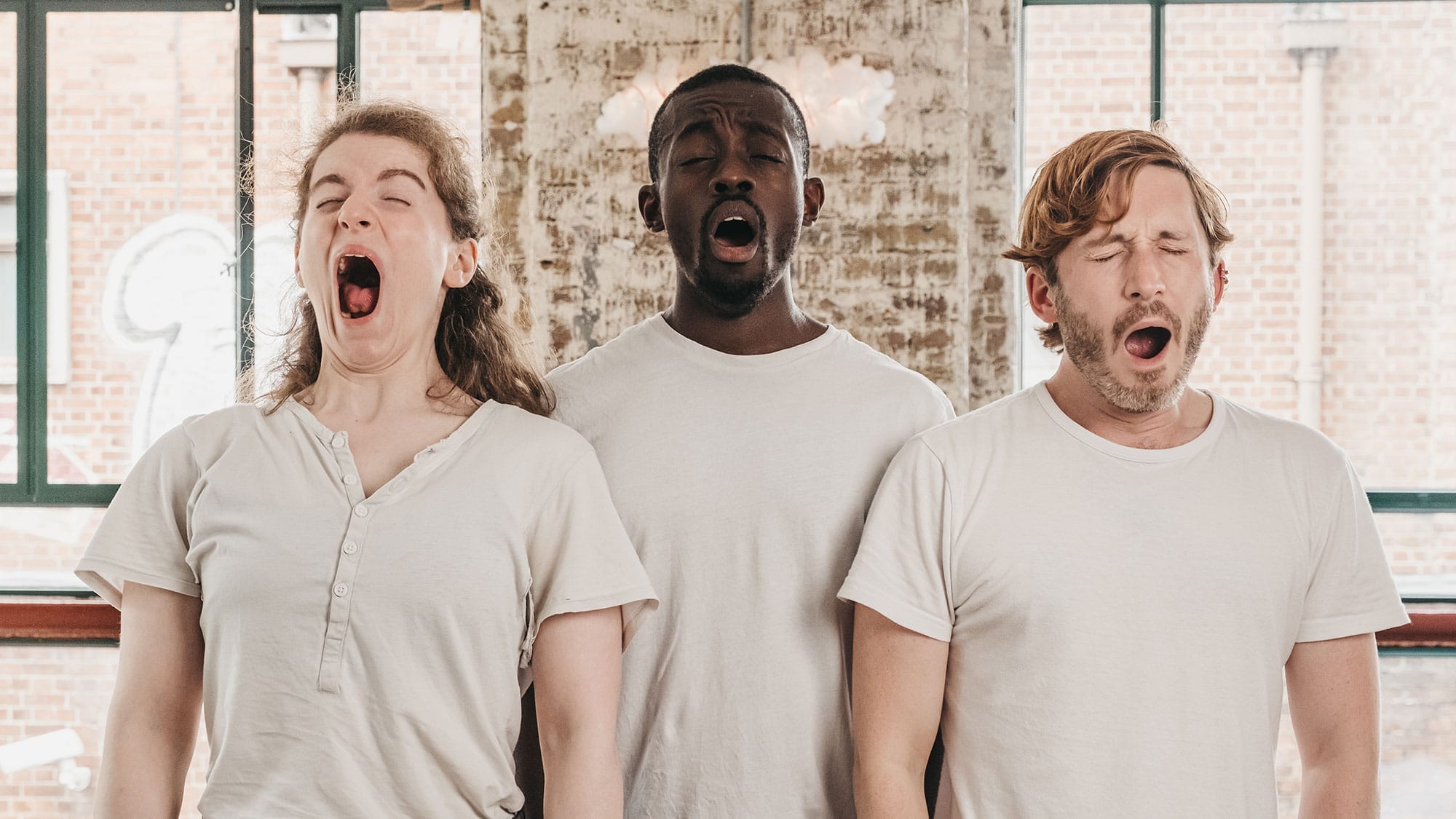 Three people stand wearing beige t-shirts all yawning.
