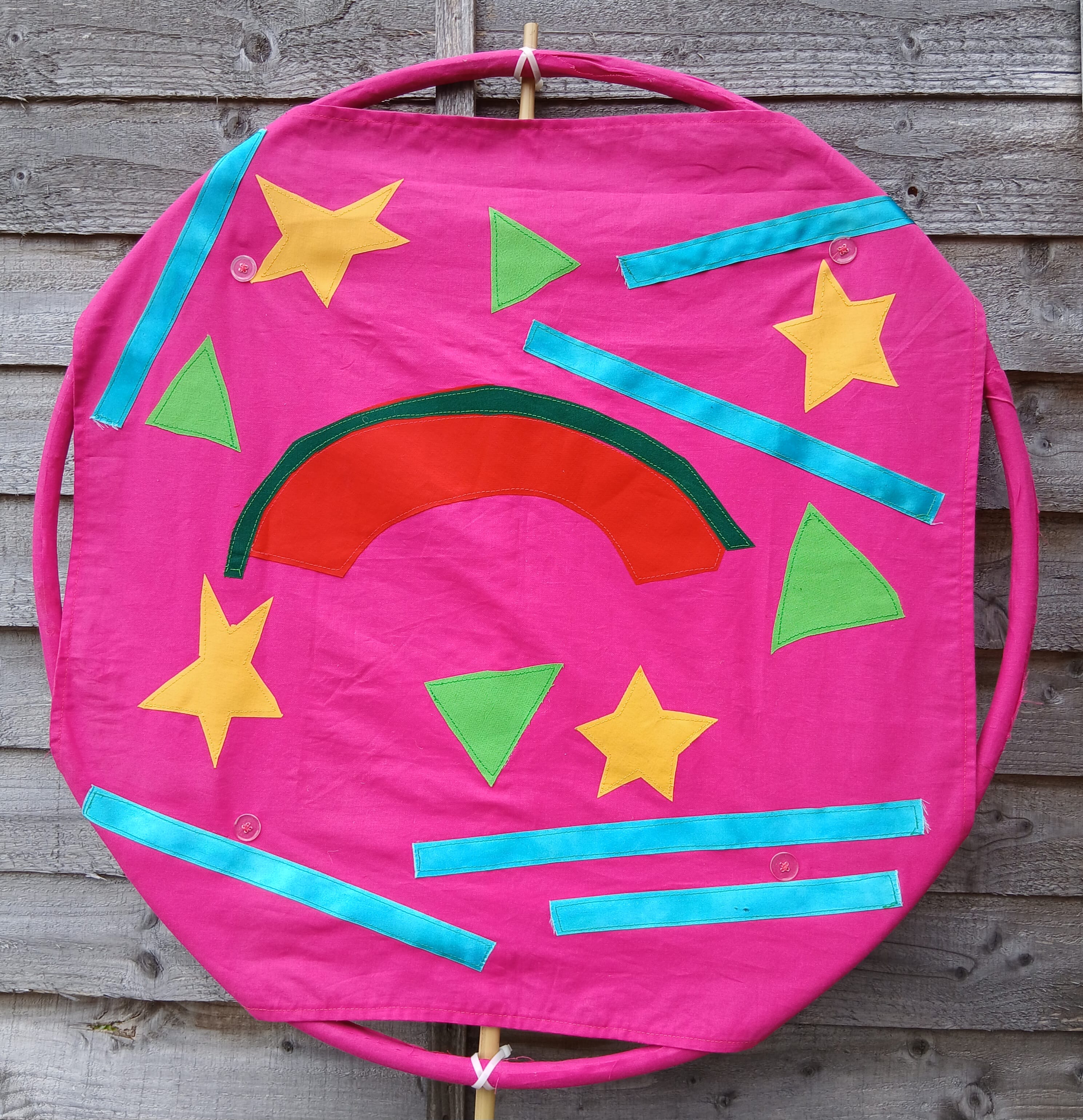 a photo of a bright pink fabric on a round frame with variously shaped colourful fabrics added on with applique method