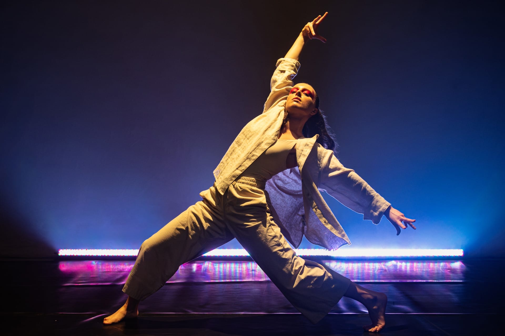 A female dancer wearing white linen trousers and shirt on a stage lit in purple