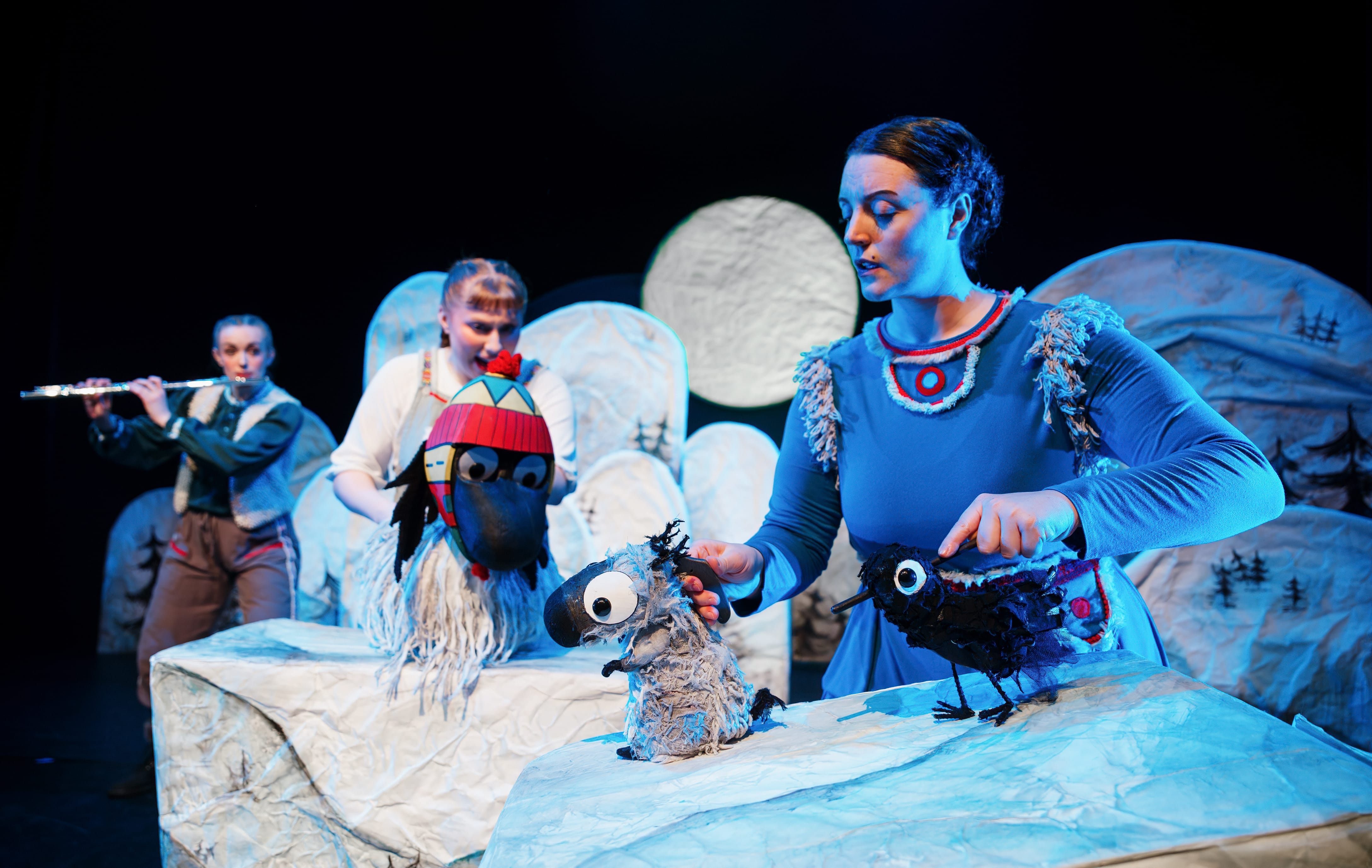three actors on a stage with a set of snowy hills handle three puppets, a yak, a bird and a marmot, with one actor playing the flute in the background