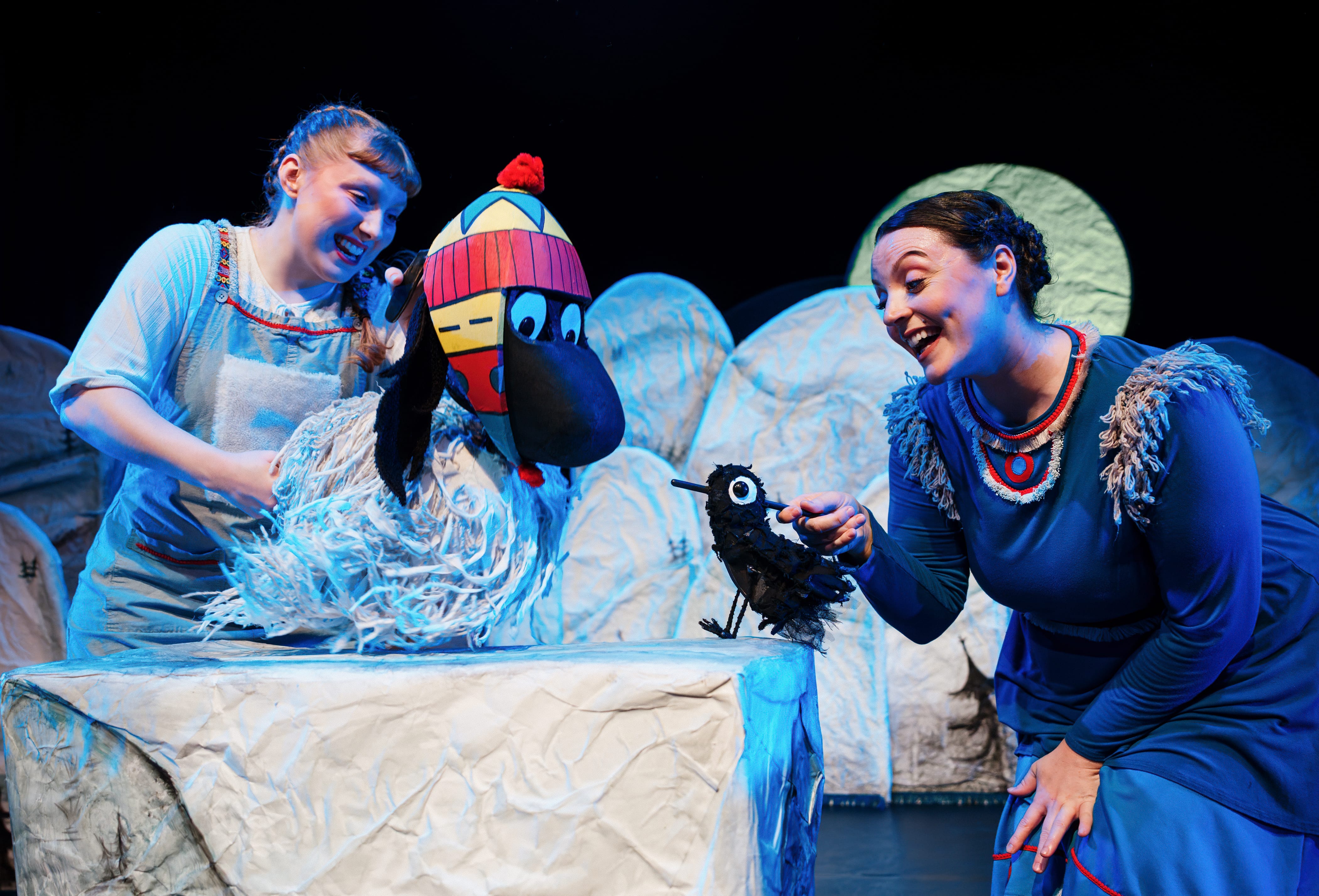 on a set with white snowy hills two actors handle a puppet each - one is a yak wearing a hat, speaking to a little black bird