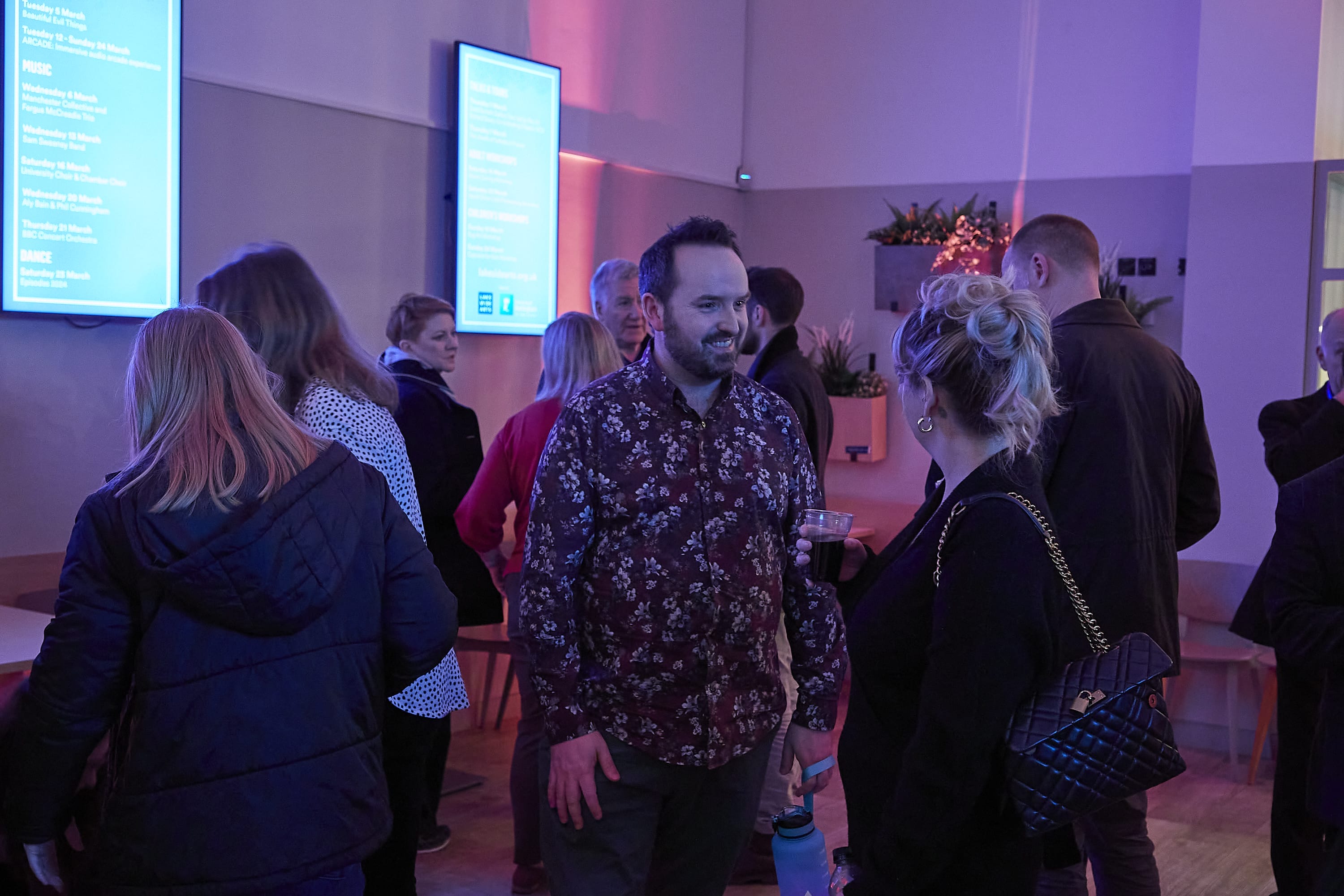 A group of people stand around chatting in a cafe space, under purple lights.