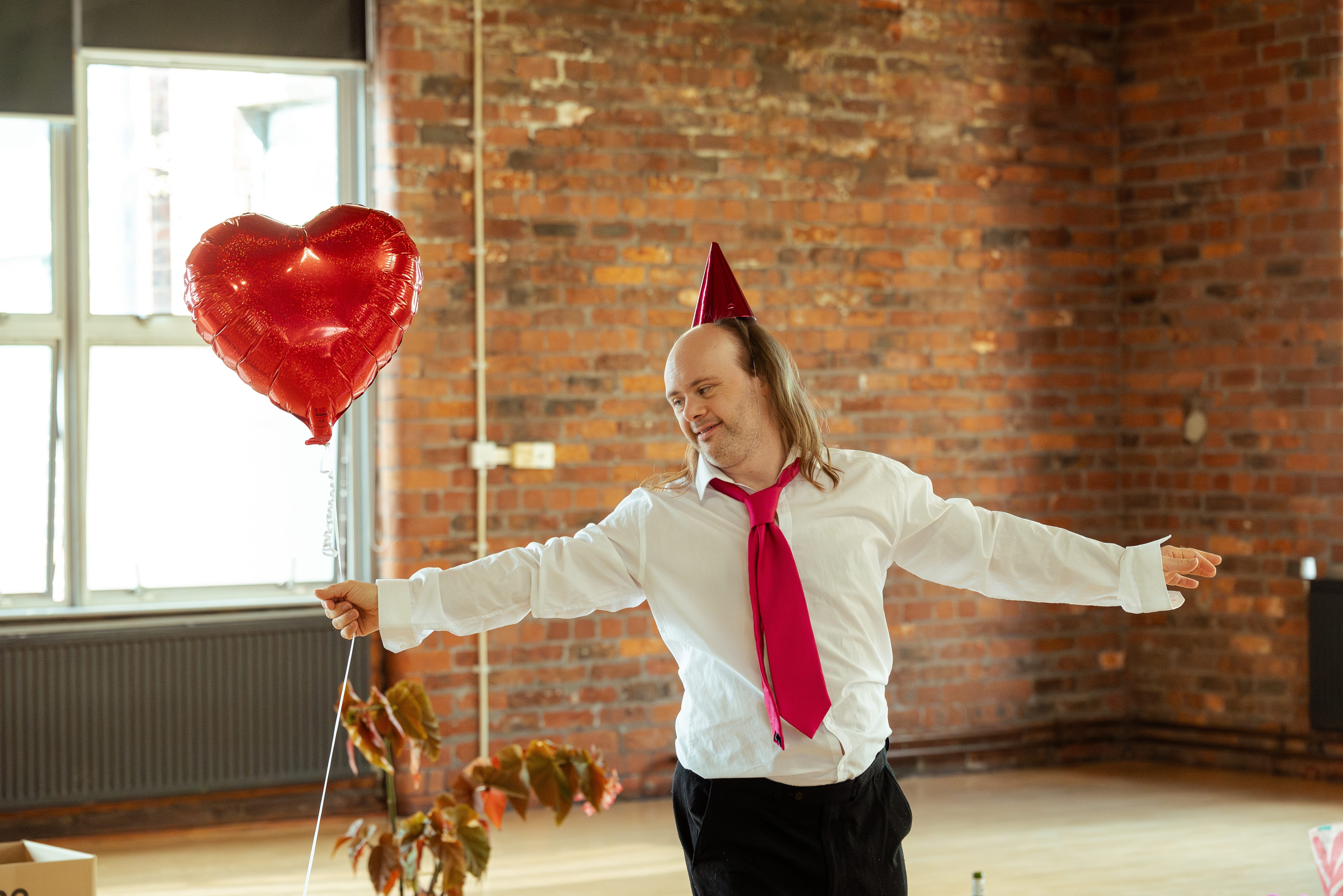 Dancer Nick McKerrow, a white male, wearing a white shirt, red tie and holding a red heart shaped balloon, stretches his arms wide in a brick walled rehearsal space.