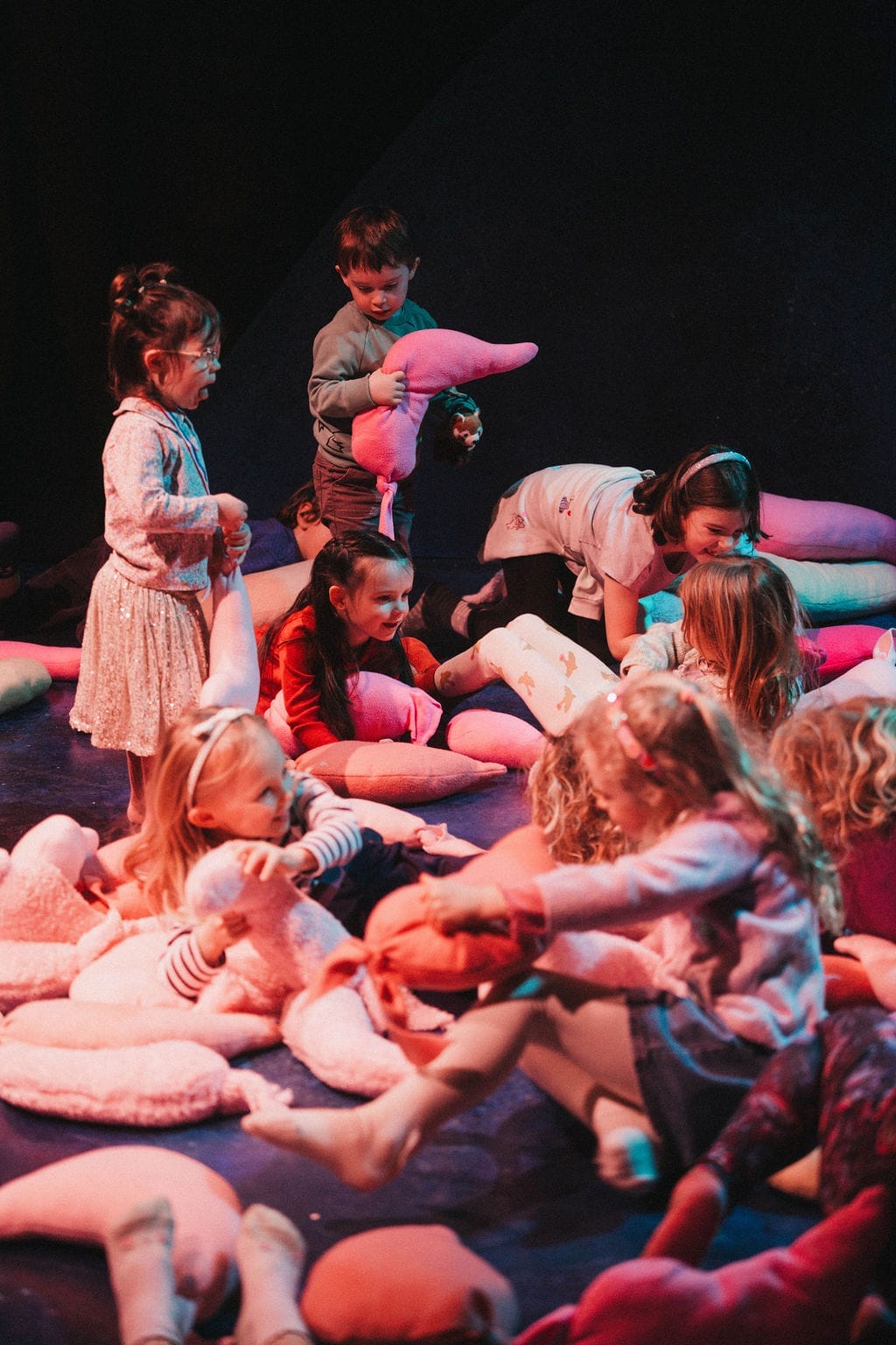 A group of children play with cushions on a dark floor.