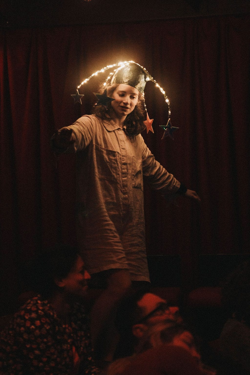 A lady with brown curly hair, wears a hat decorated with lights and stars, in a darkened space.
