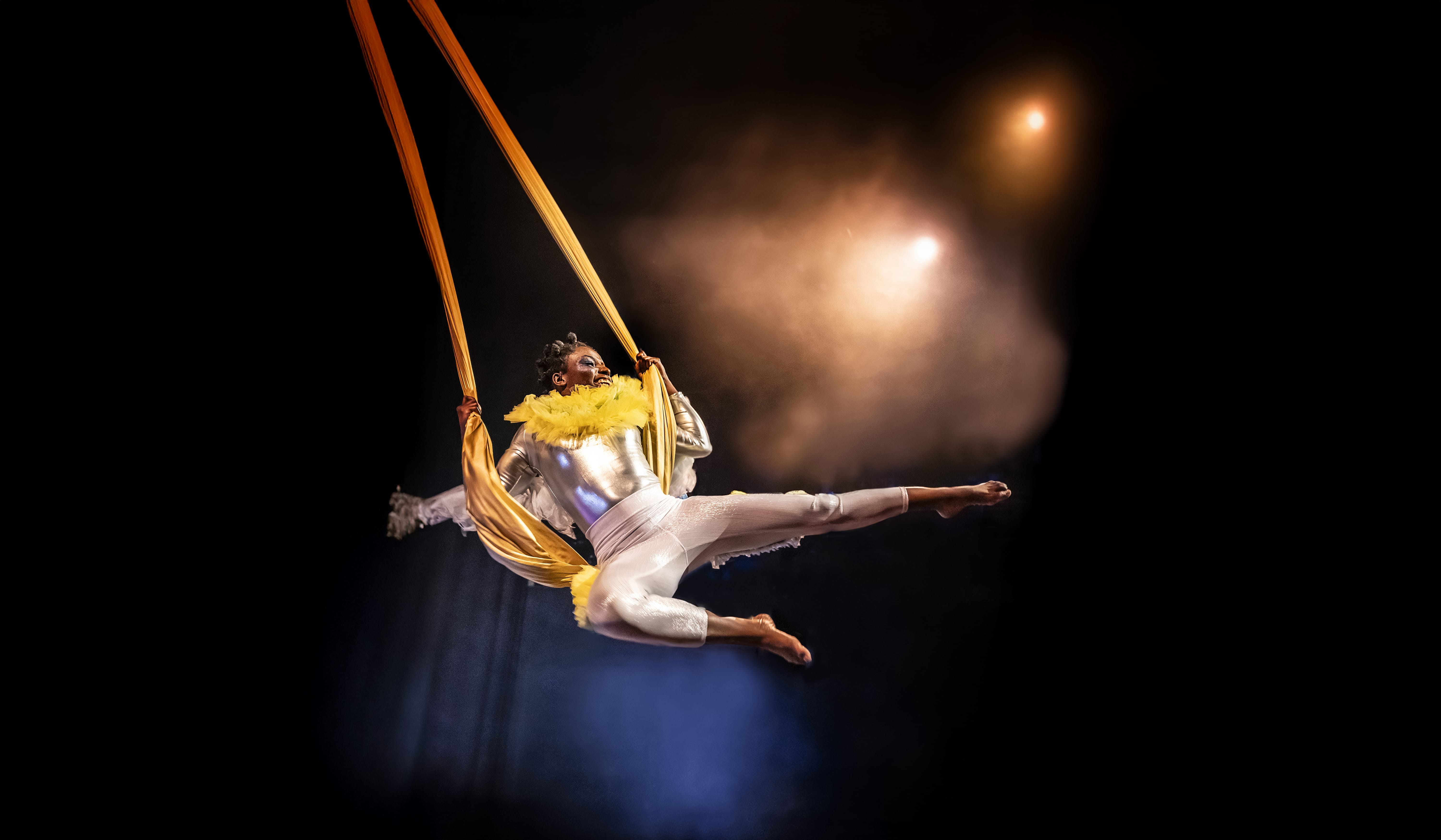 a black woman dressed in shiny lycra holds onto a silk swing and flies in the air across a dark stage