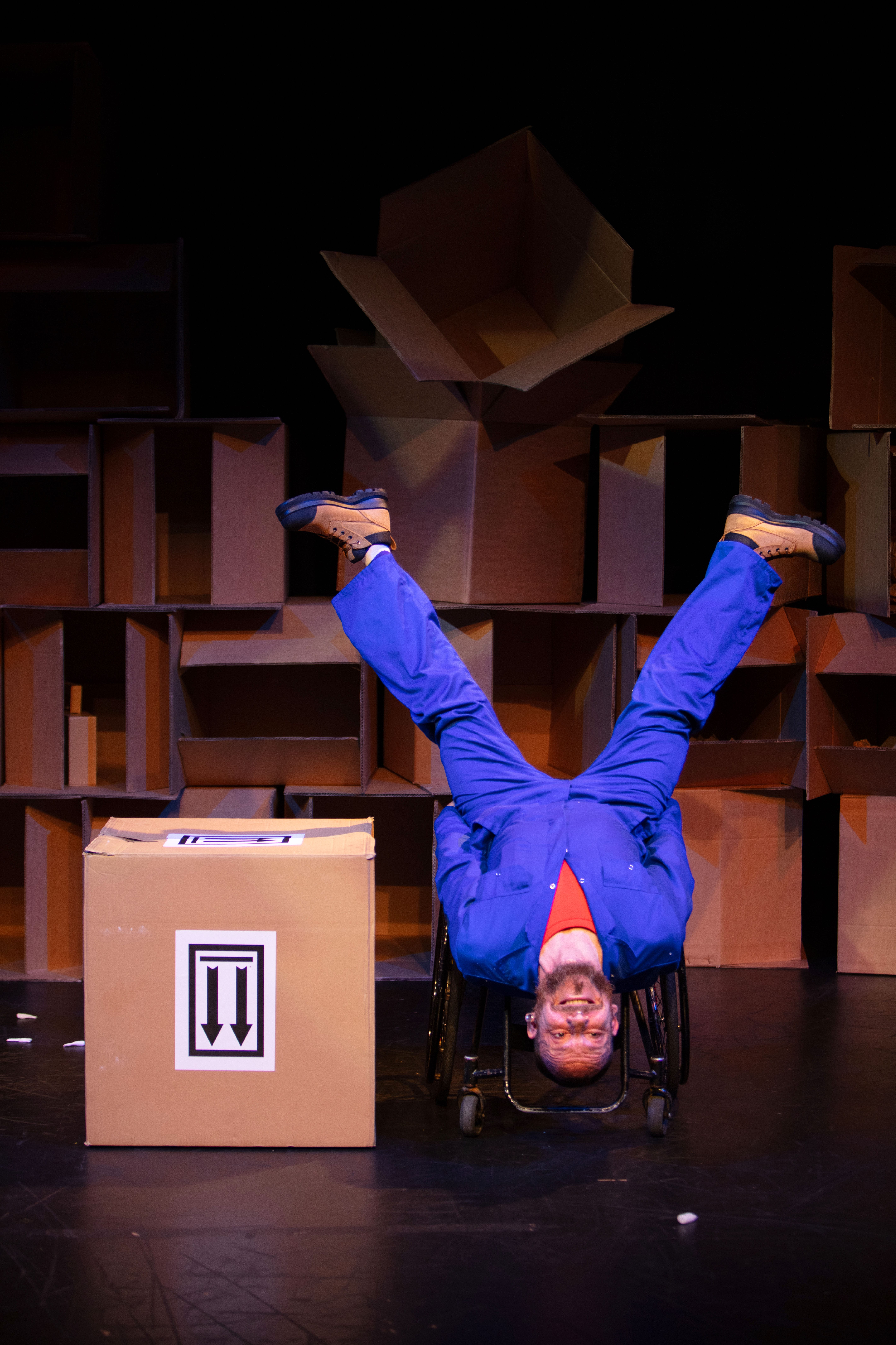 A man in a blue boiler suit is upside-down with legs spread wide, in a wheelchair, next to a cardboard box.