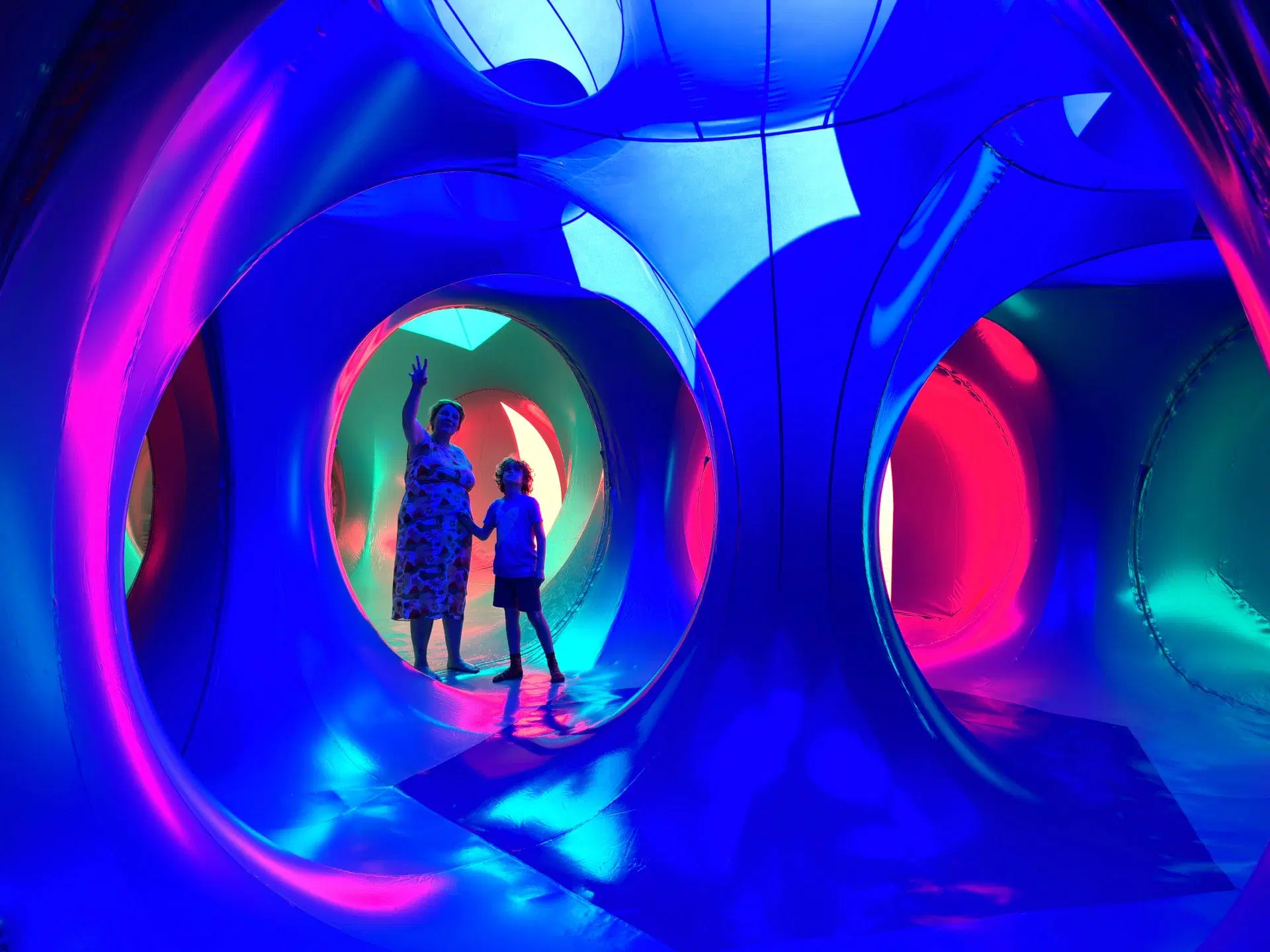 A lady and small boy hold hands as they look up in a tunnelled structure of the luminarium, bathes in blue light.
