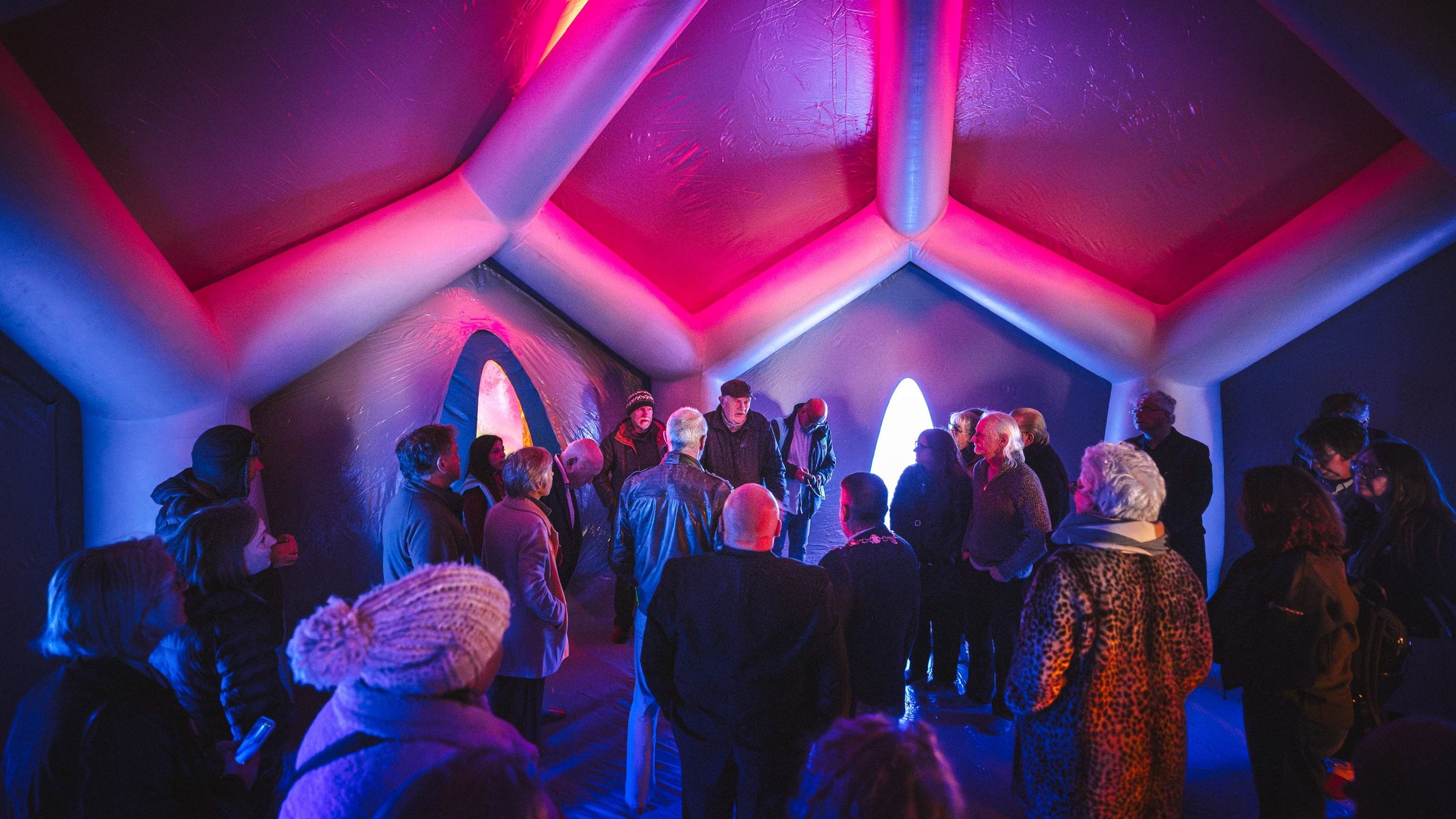 A large group of people stand under the pink light of the luminarium.