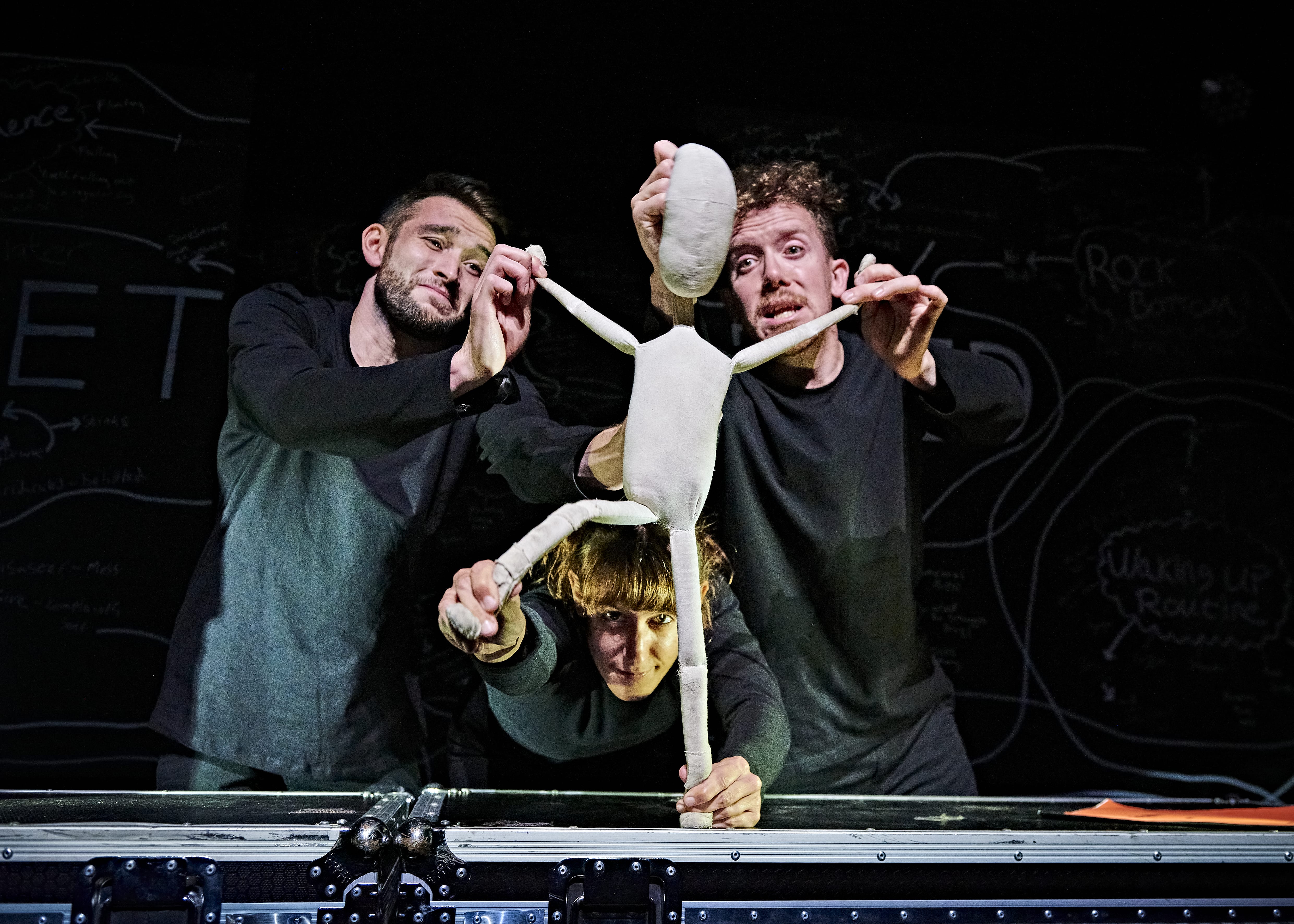 Three puppeteers, dressed in black, hold a white human-shaped puppet up to the camera.