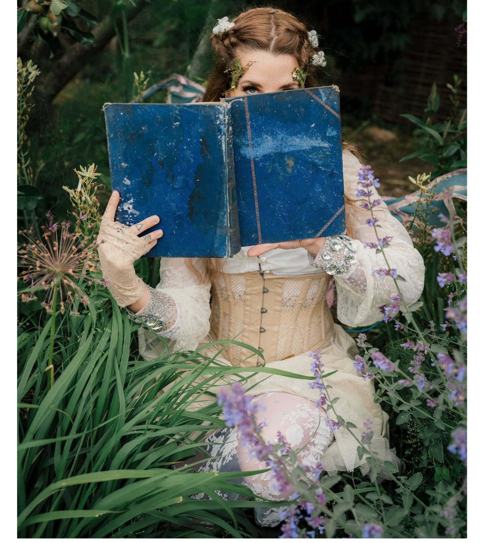A woman in historic dress, sits among wild flowers and peers from behind a blue book.