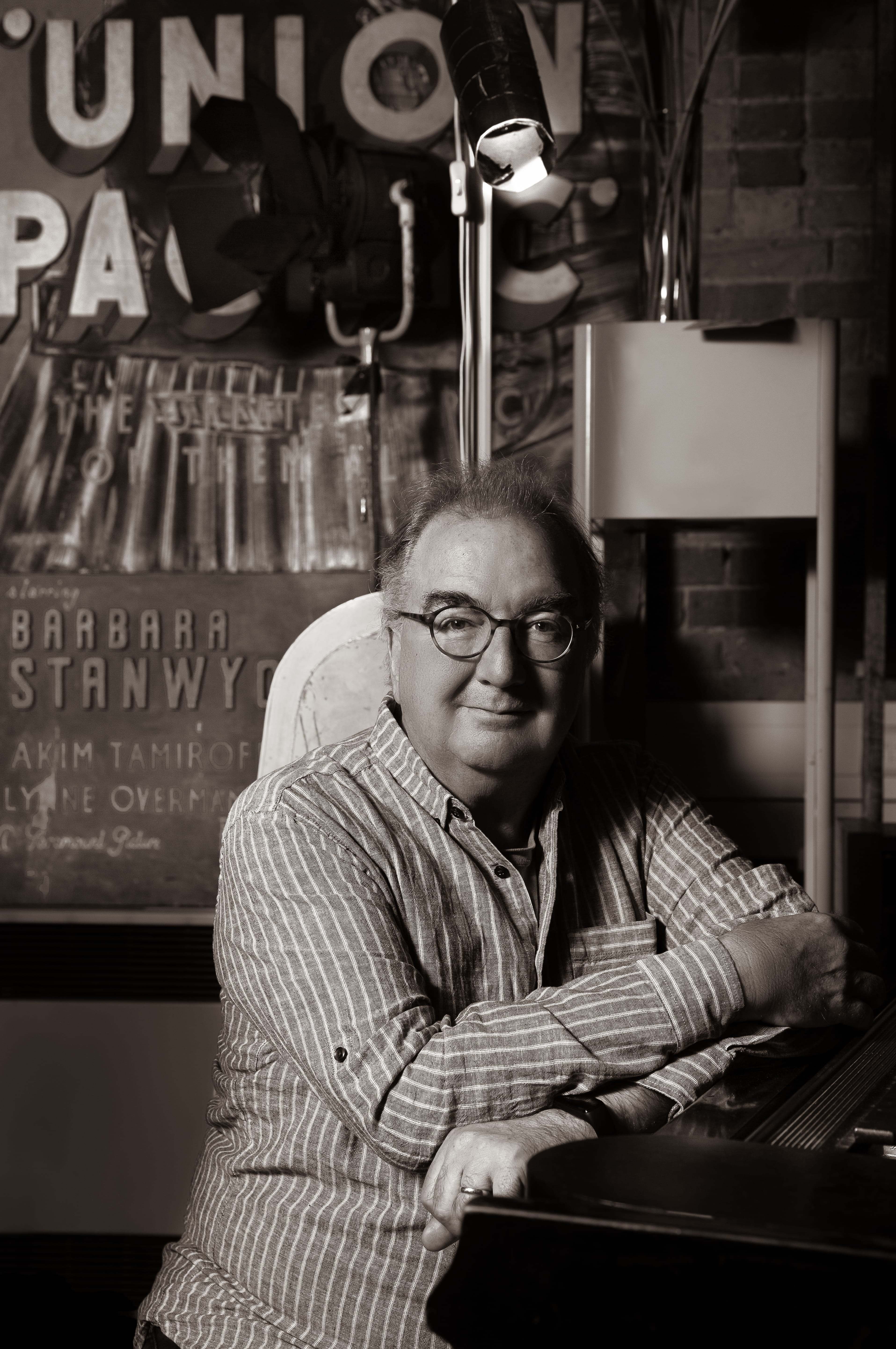 Neil Brand, a man wearing glasses and a striped shirt, sits at a piano. In a black and white photograph.