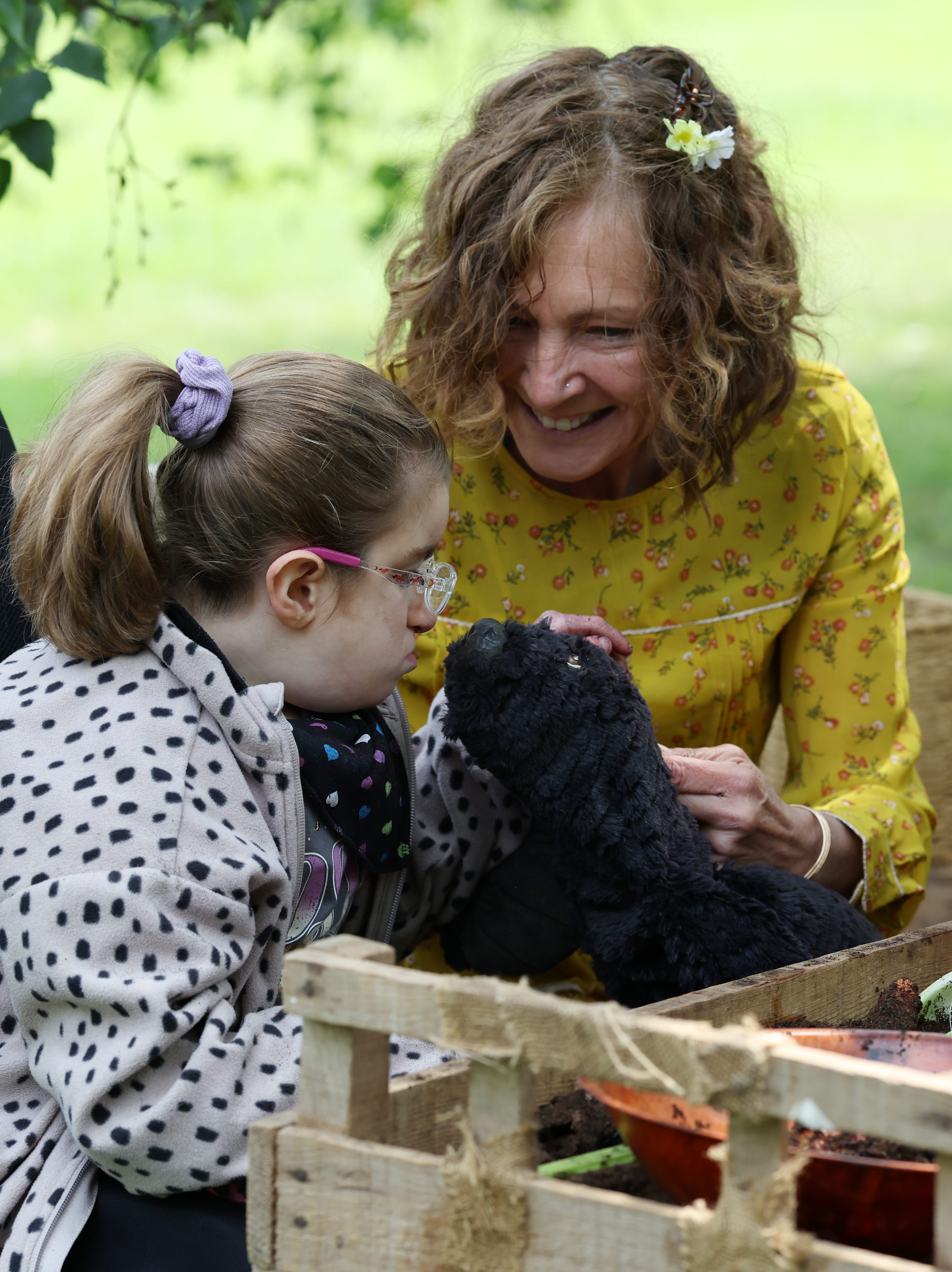A lady with curly hair entertains a girl in a black and white spotted jumper, with a puppet.