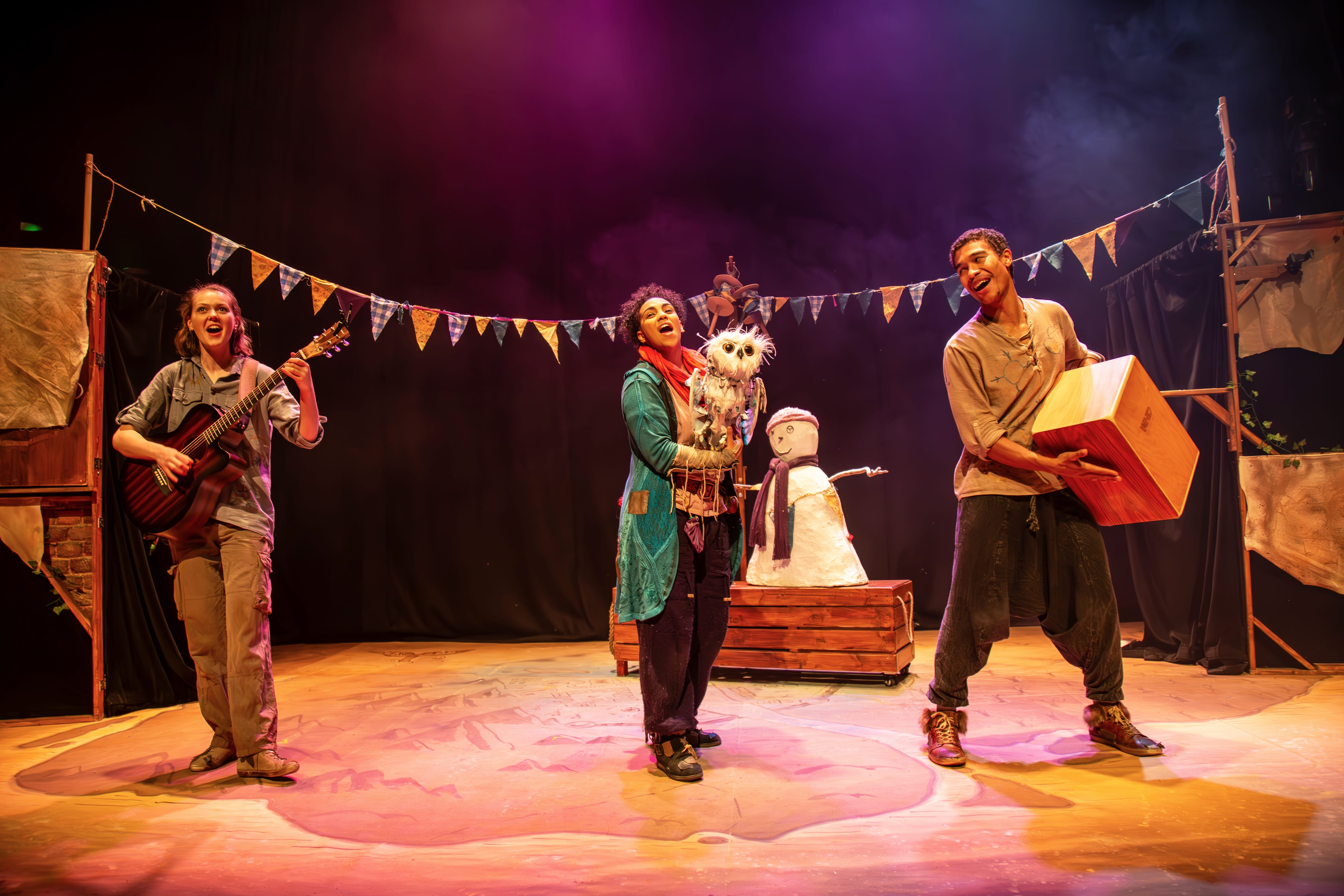 Three actors on stage with a bunting in the background, one holding an owl puppet, the other two playing on musical instruments