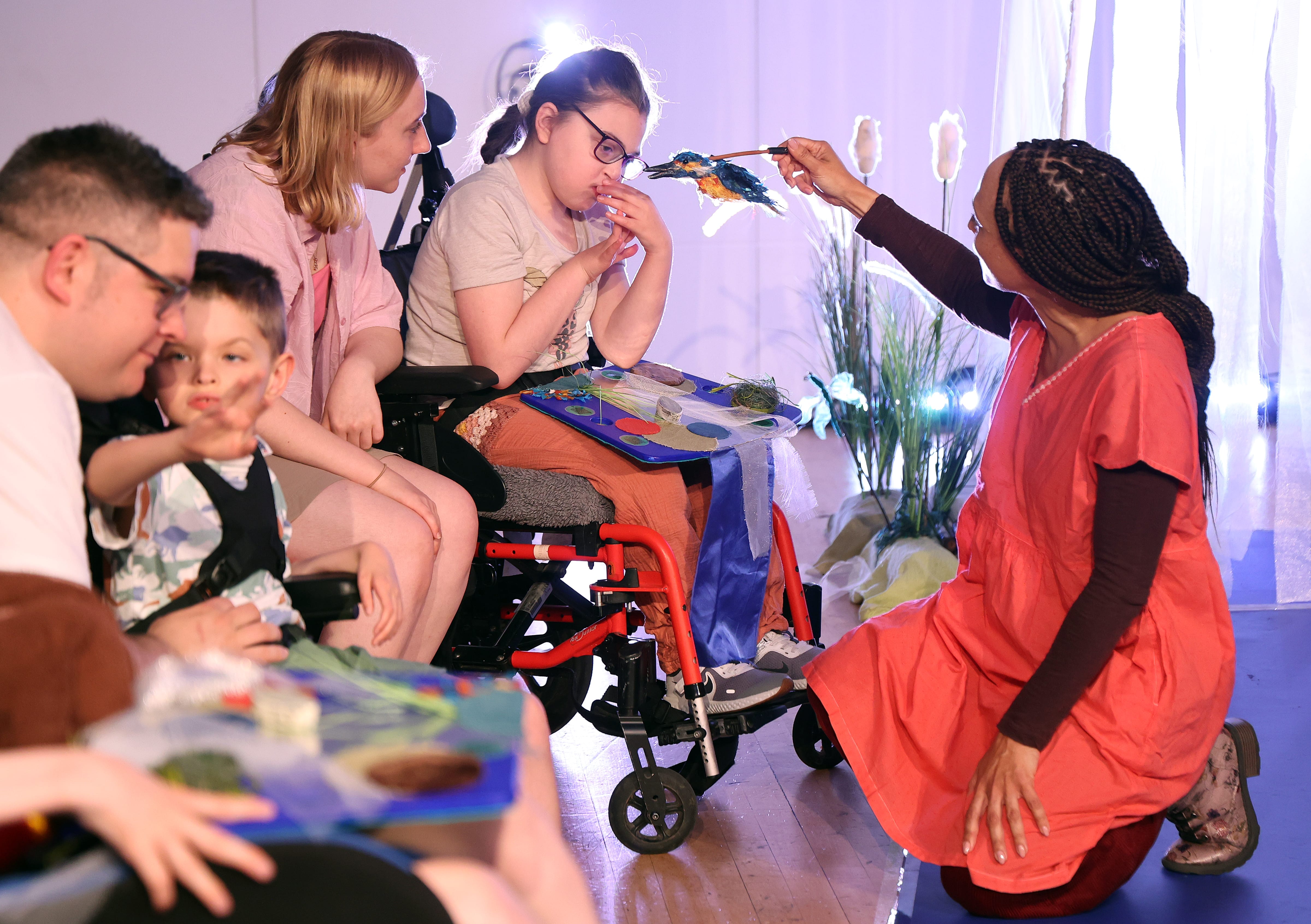 A group of adults and children, enjoy interactive theatre. A girl wearing glasses, sits in a red wheelchair enjoying a kingfisher puppet.