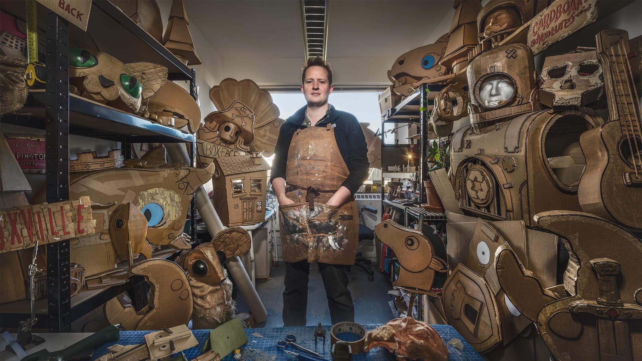 A man wearing an apron, stands in a workshop surrounded by cardboard creations.