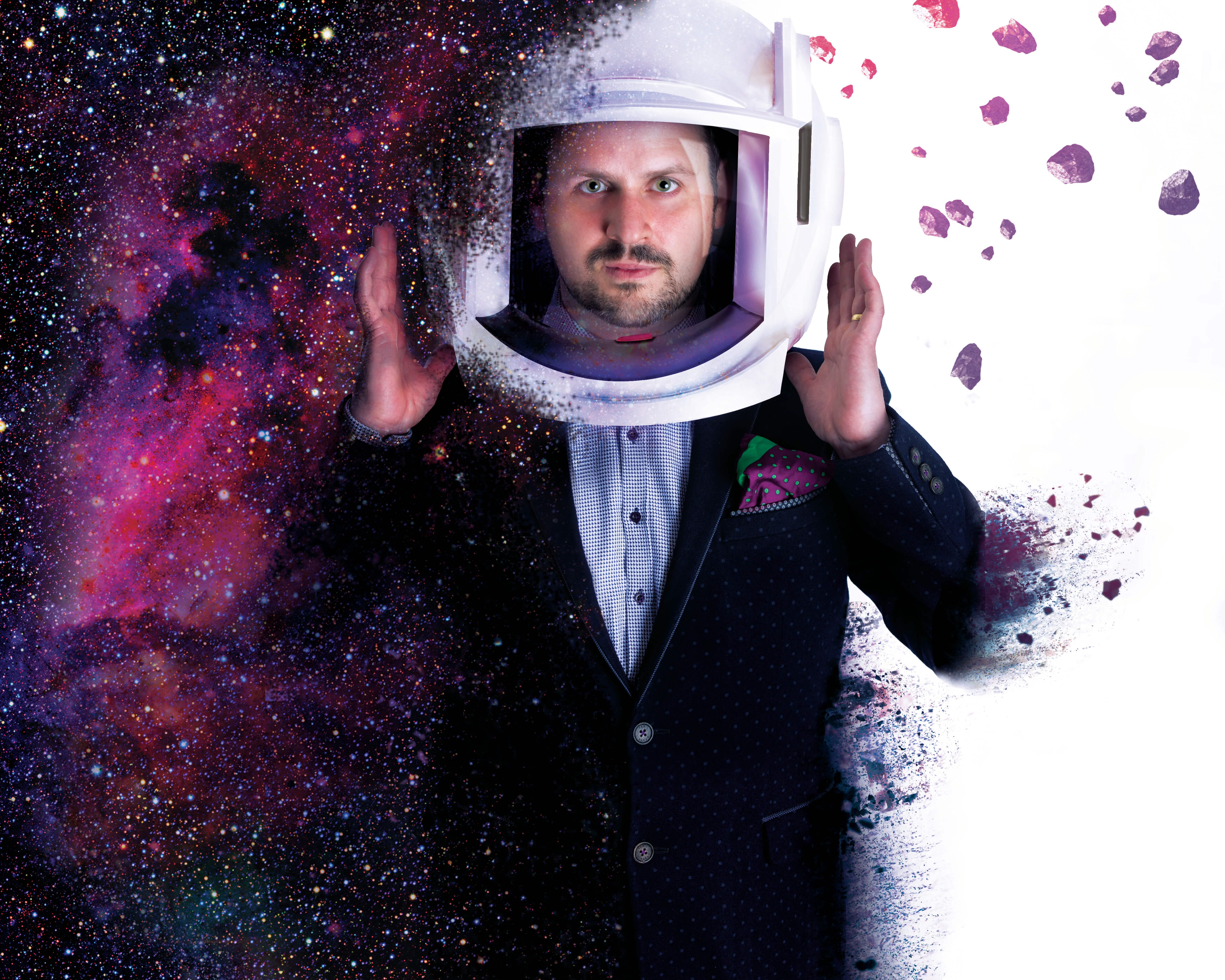 A man wearing a space helmet and sports jacket holds his hands in the air, either side of the helmet.