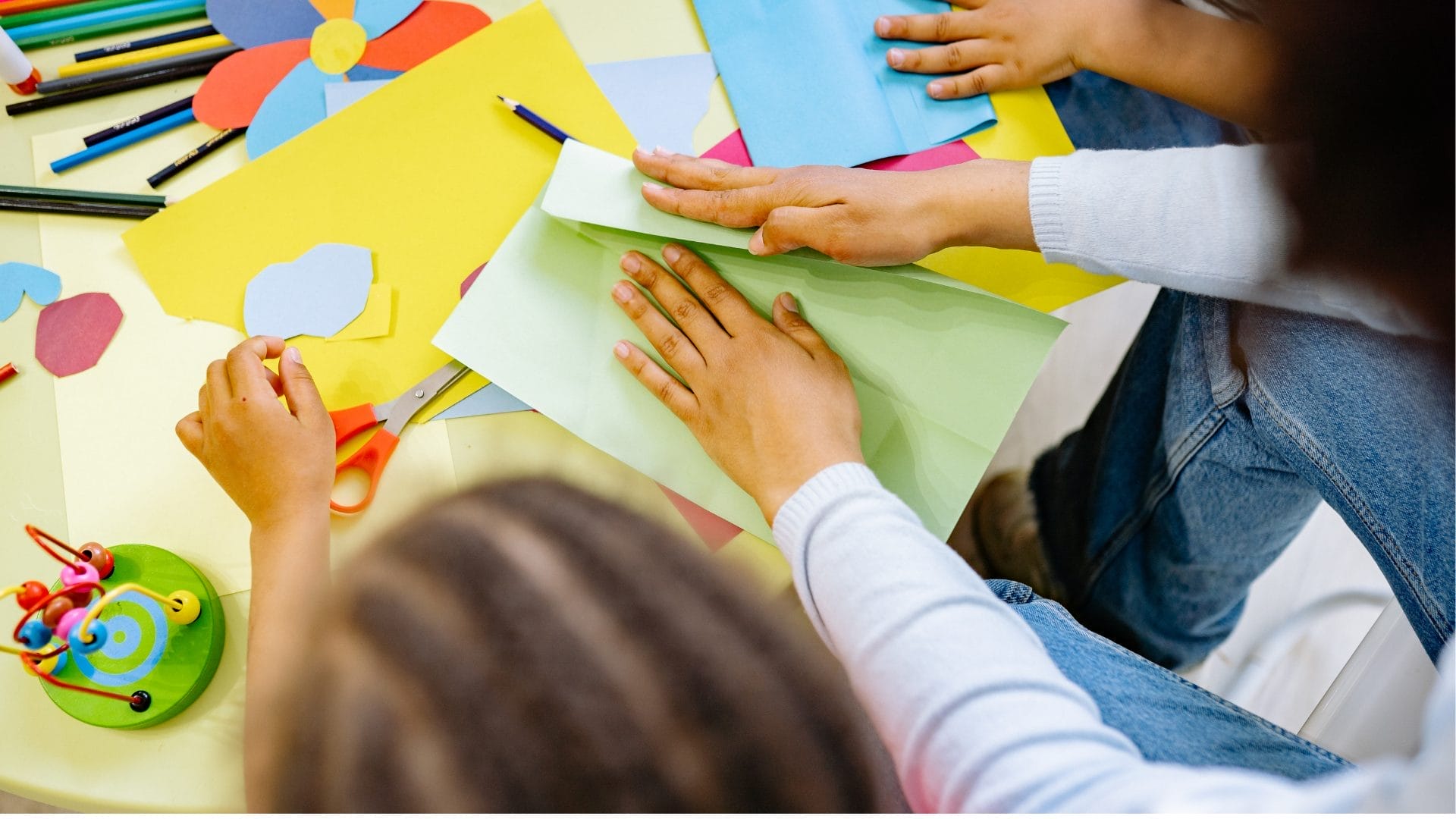 Children are crafting at a table, you can see small hands folding a green piece of paper.