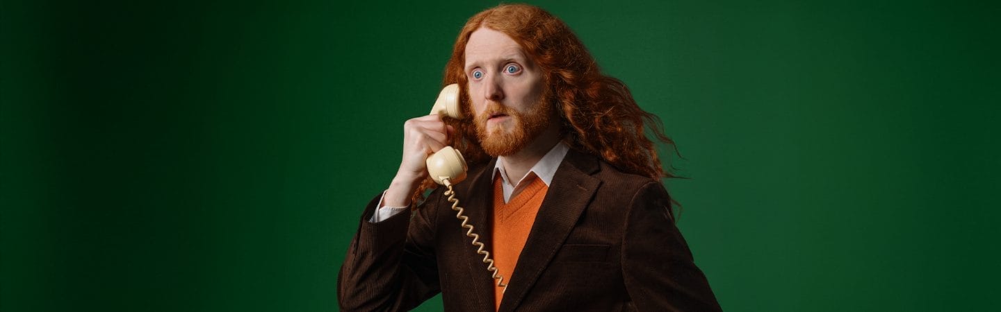 A man with long, flowing ginger hair - stares into the distances whilst holding a phone to his ear.